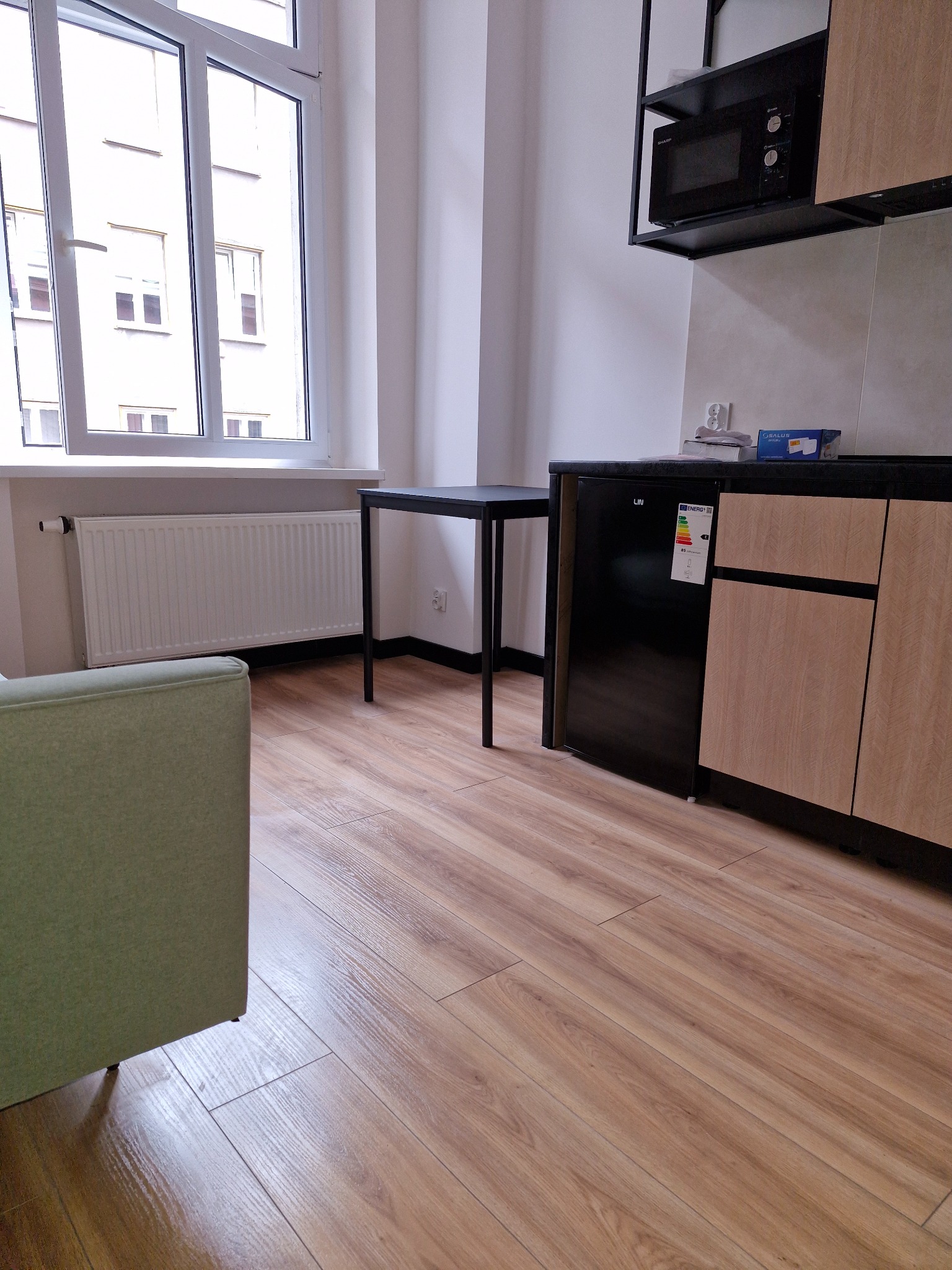 Nowoczesna kawalerka z drewnianą podłogą, minimalistycznym aneksem kuchennym z mikrofalówką i małą lodówką, grzejnikiem pod oknem i fragmentem zielonej sofy.