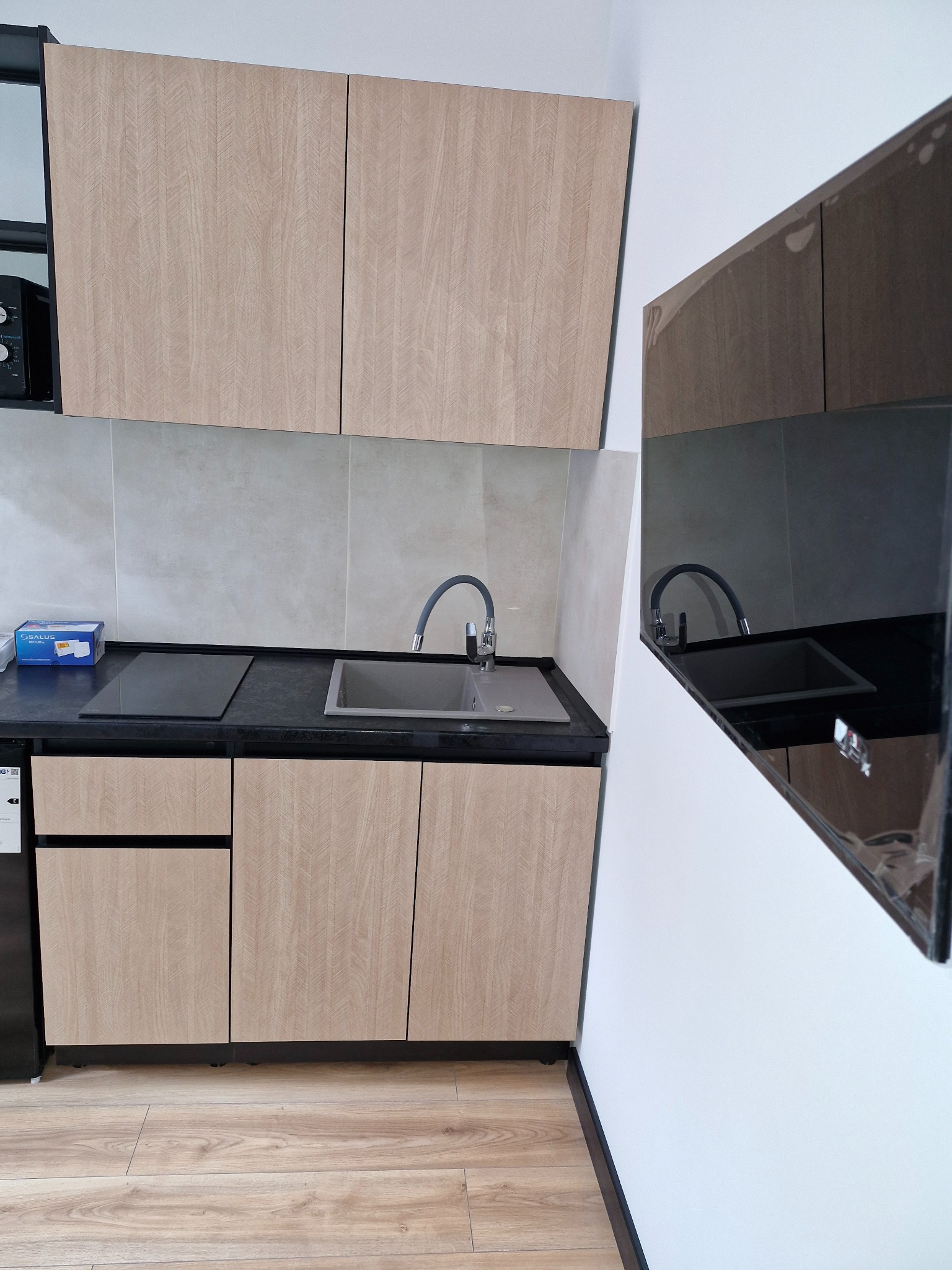 Nowoczesna, jasna kuchnia z drewnopodobnymi frontami szafek, szarym blatem i zlewozmywakiem oraz płytą grzewczą. W odbiciu na ścianie widoczny fragment tego samego blatu i zlewu.