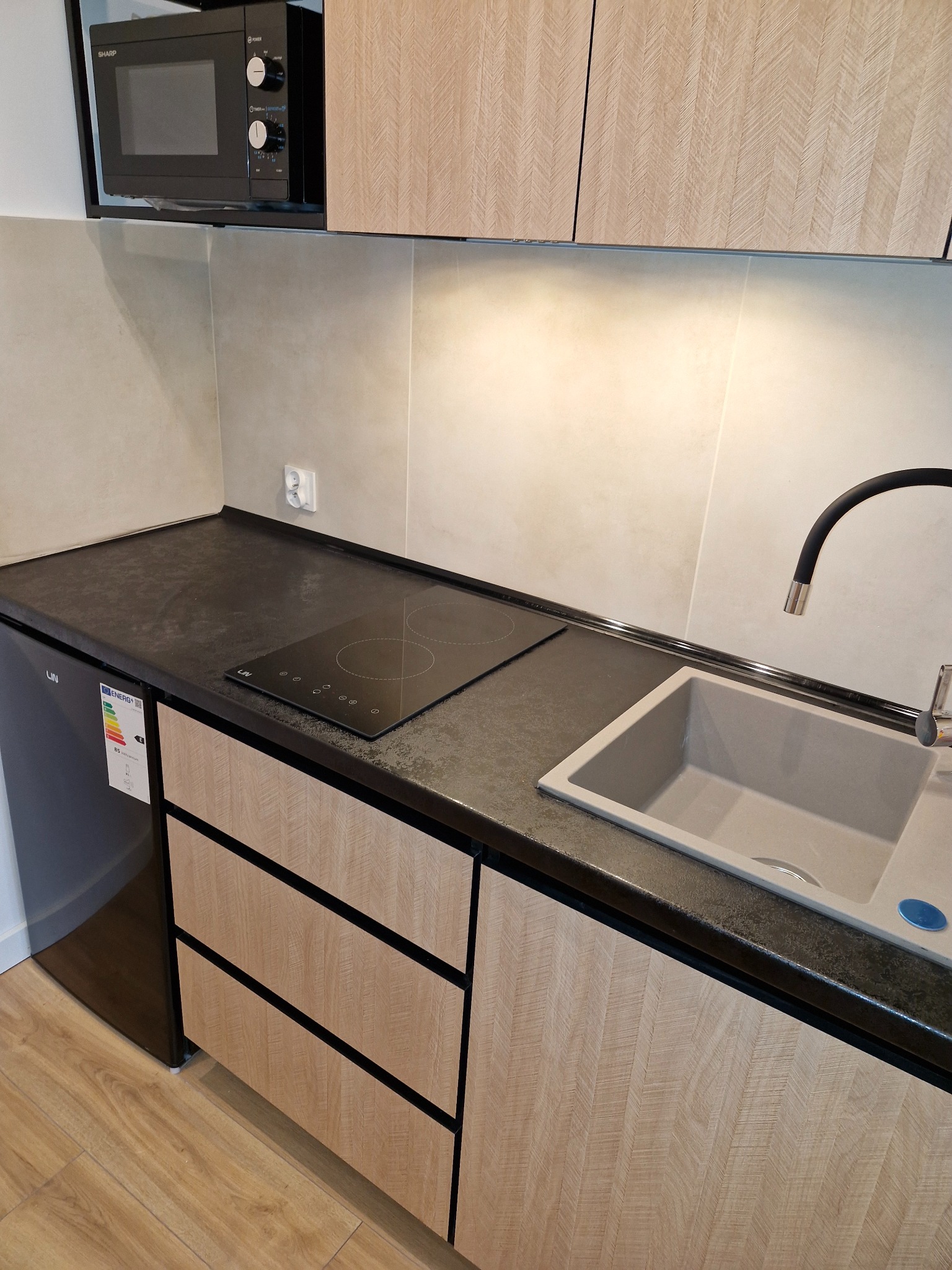 Nowoczesna kuchnia z drewnopodobnymi frontami szafek, czarnym blatem, płytą indukcyjną i szarym zlewem; widoczny również czarny mikrofalówka Sharp.