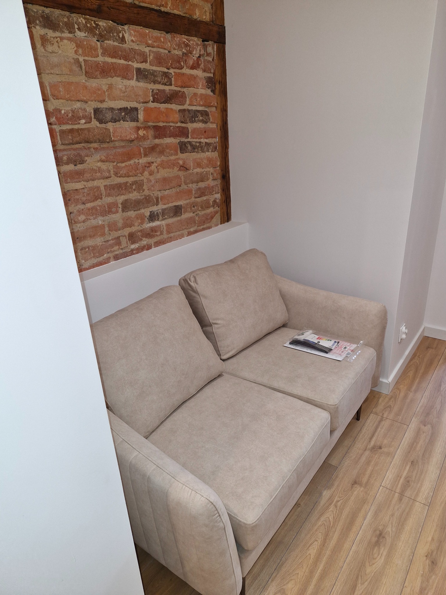 Beżowa sofa dwuosobowa z poduszkami na tle ściany z cegły i drewnianej belki stropowej, na siedzisku leżą dokumenty w folii, podłoga drewniana.