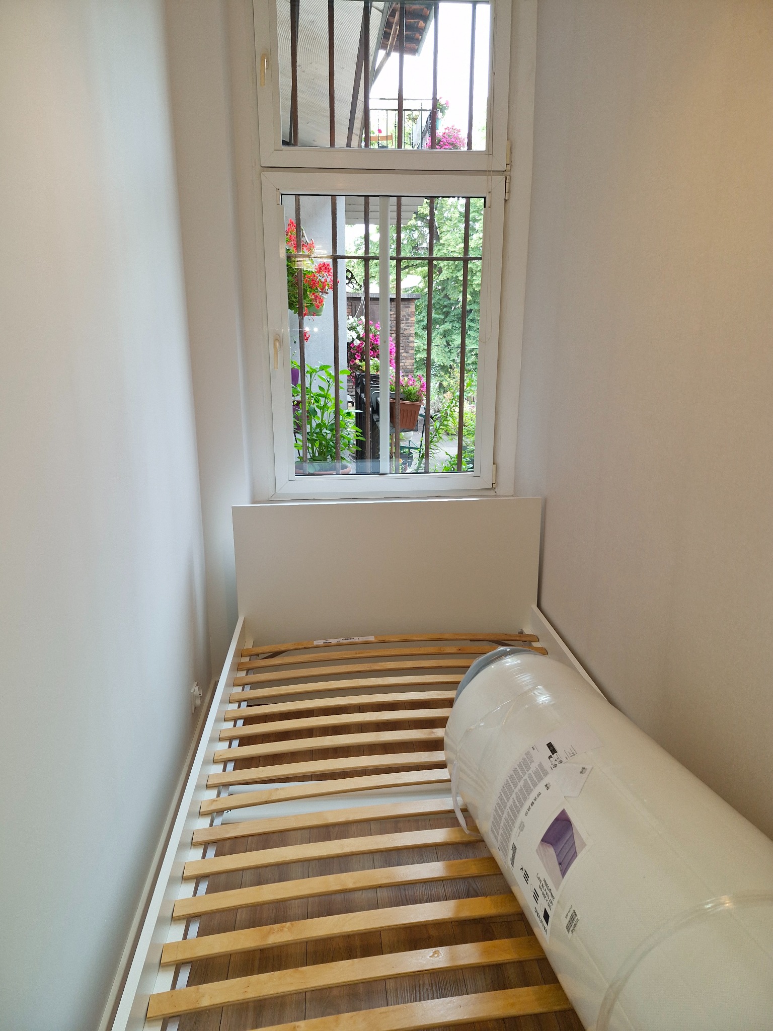 Rozpakowany materac w folii leży na drewnianym stelażu łóżka w pokoju z oknem z kratami, za którym widać rośliny i kwiaty.