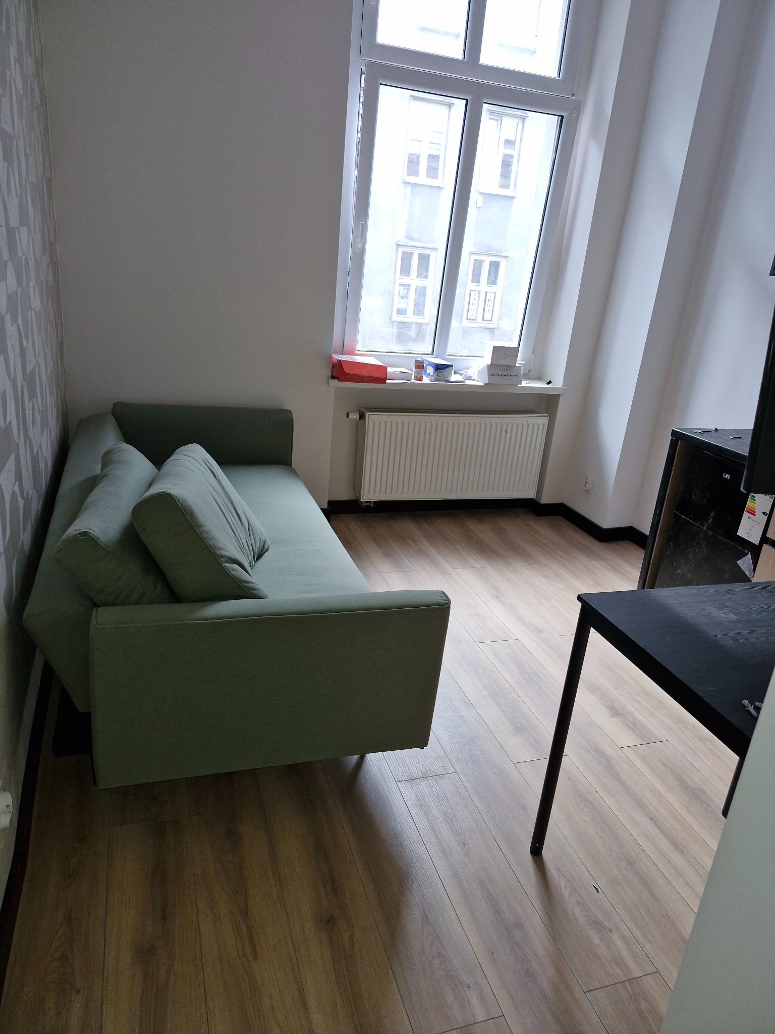 Jasnozielona rozkładana sofa z poduszkami w pokoju z drewnianą podłogą i grzejnikiem pod oknem.