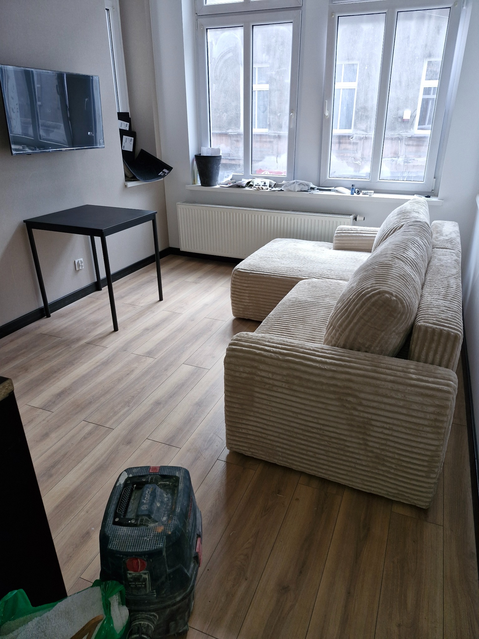 Jasna, modułowa sofa z prążkowaną tapicerką w pokoju z panelami podłogowymi, widoczne duże okno i odkurzacz przemysłowy.