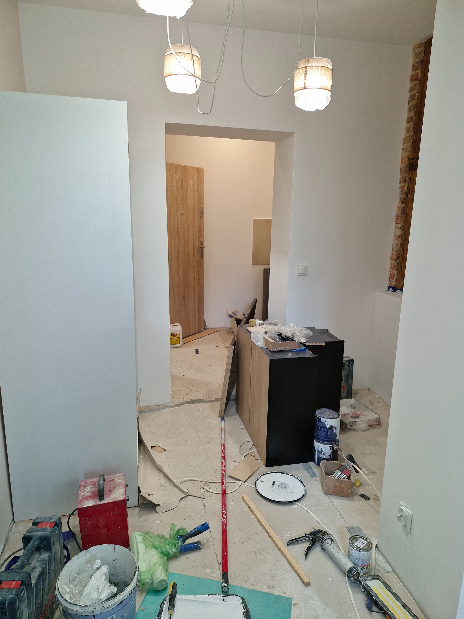 Wnętrze w trakcie prac wykończeniowych: białe ściany, widoczne narzędzia malarskie, wiadro z farbą, puszki z farbami, listwy przypodłogowe, lampy wiszące i fragment odsłoniętej cegły.