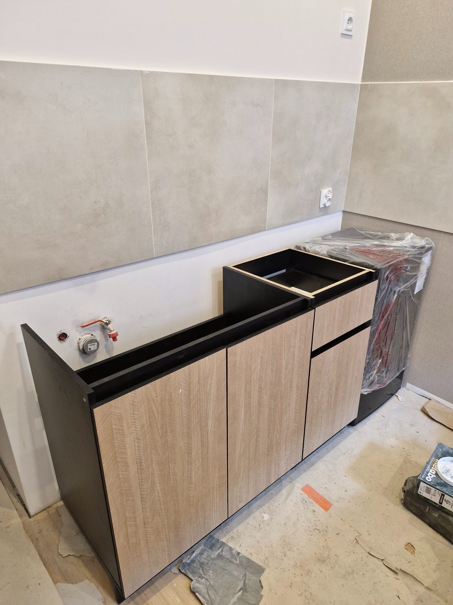 Montaż szafki pod umywalkę w łazience z szarymi płytkami, widoczne przyłącza wodne i zabezpieczona folią pralka.