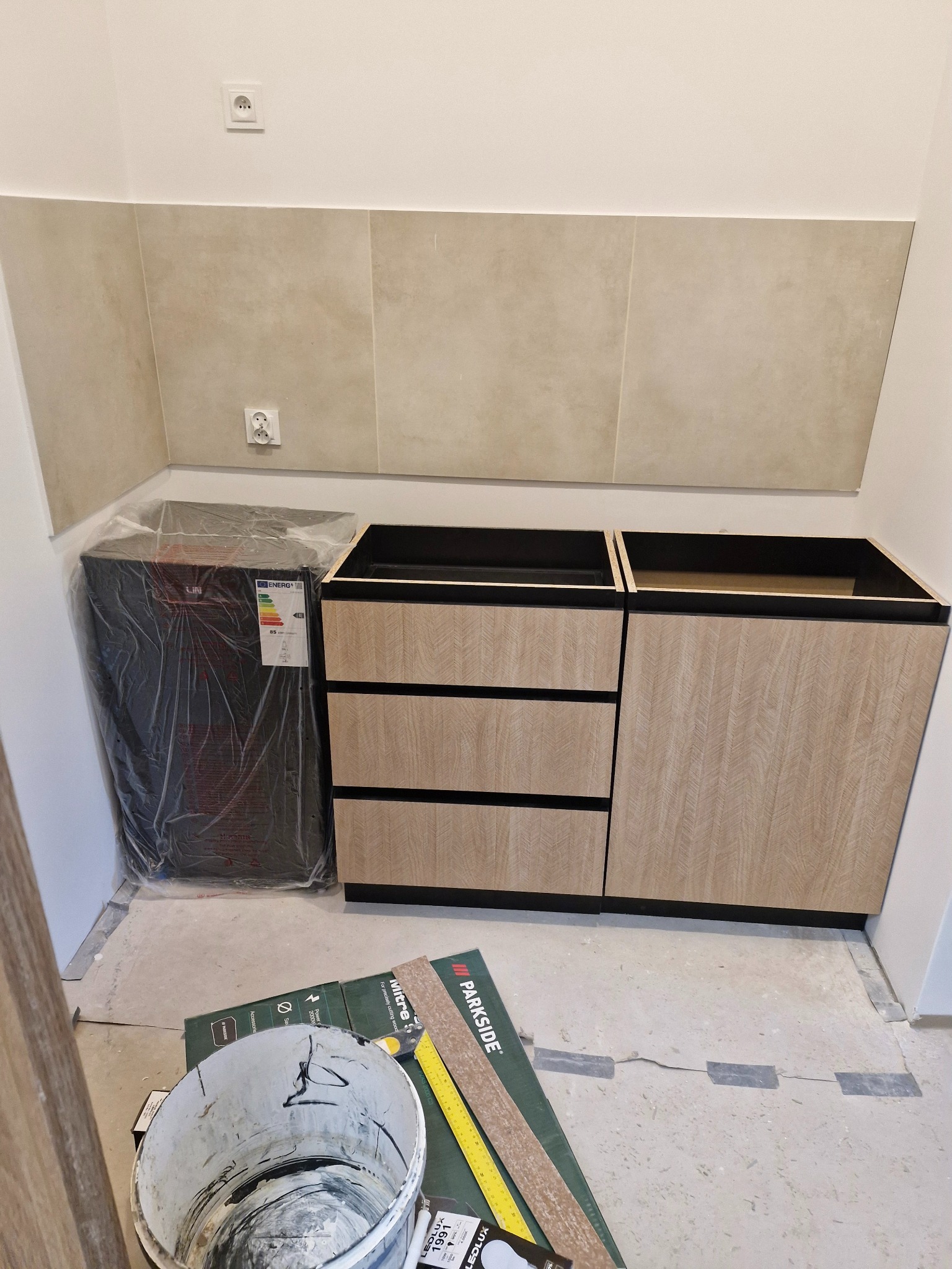 Montaż szafek kuchennych w łazience, widoczny pojemnik z resztkami farby, płytki ceramiczne na ścianie i zapakowana umywalka, etap prac wykończeniowych.