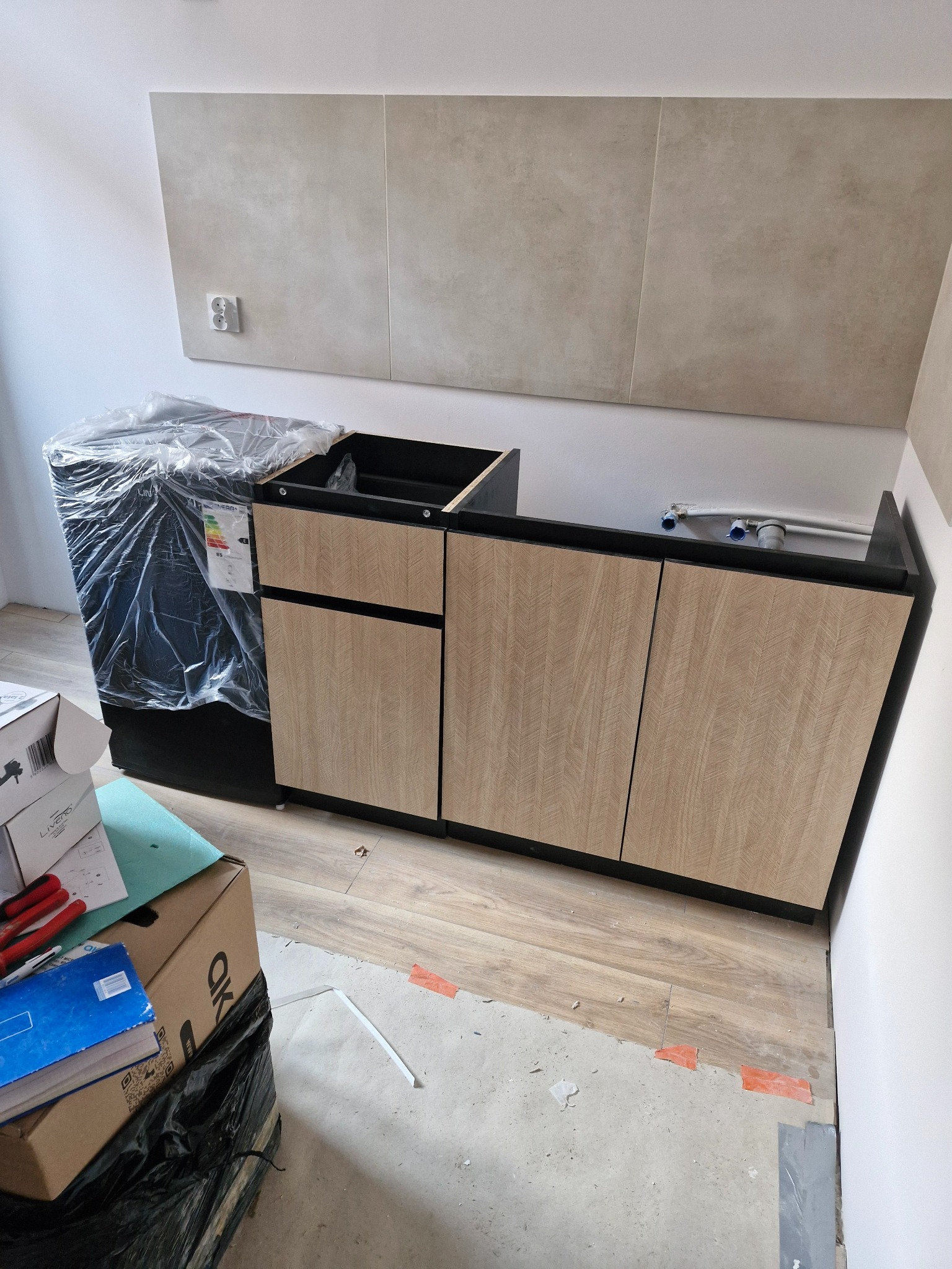 Częściowo zamontowana szafka pod umywalkę z drewnopodobnymi frontami i czarnym blatem, obok pralka w folii ochronnej, widoczne płytki na ścianie i niedokończona podłoga.