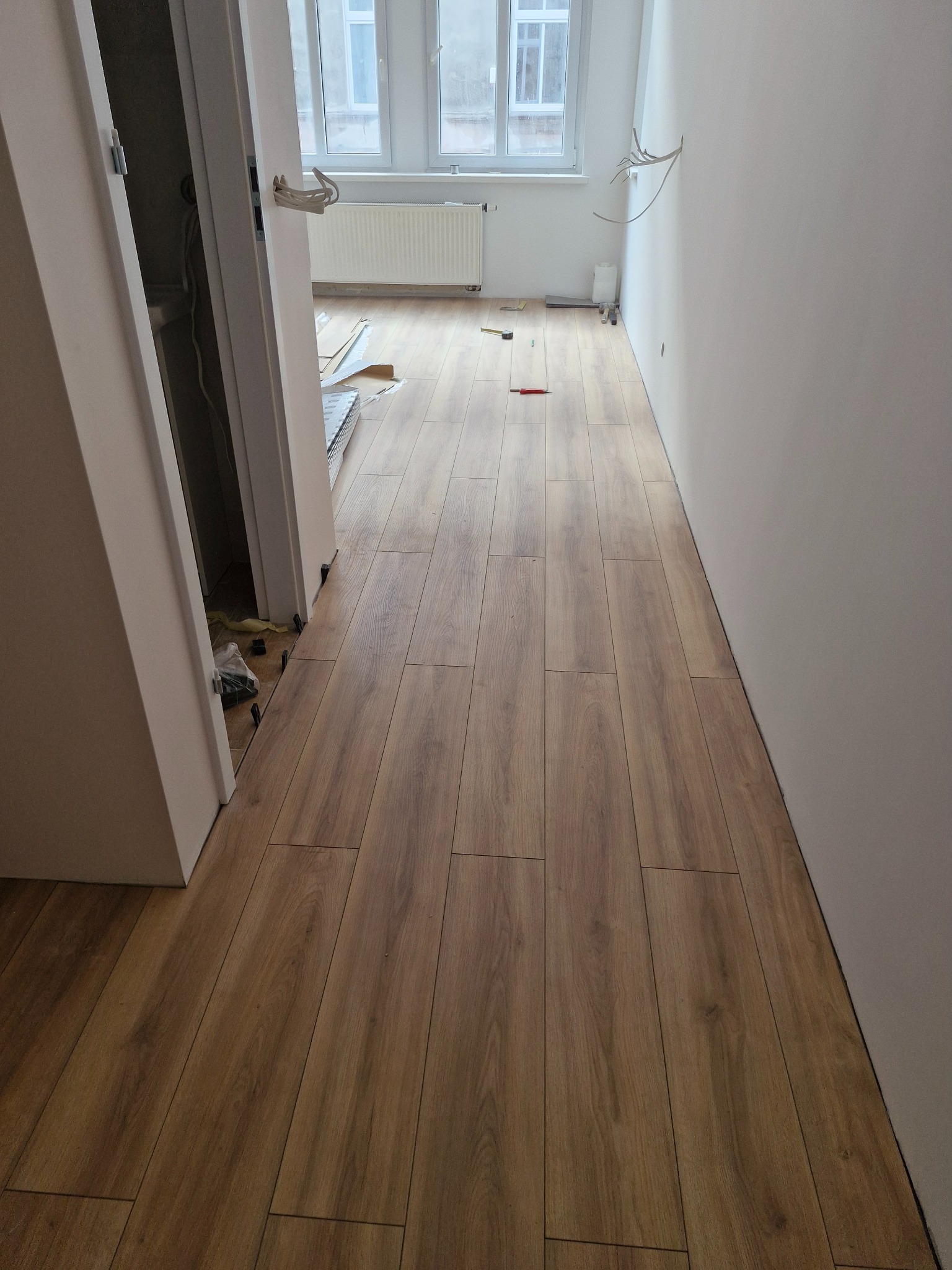 Ułożona podłoga laminowana o ciepłym odcieniu drewna w jasnym pomieszczeniu z białymi ścianami i oknem w tle, widoczne niedokończone prace przy listwach przypodłogowych oraz wystające kable.