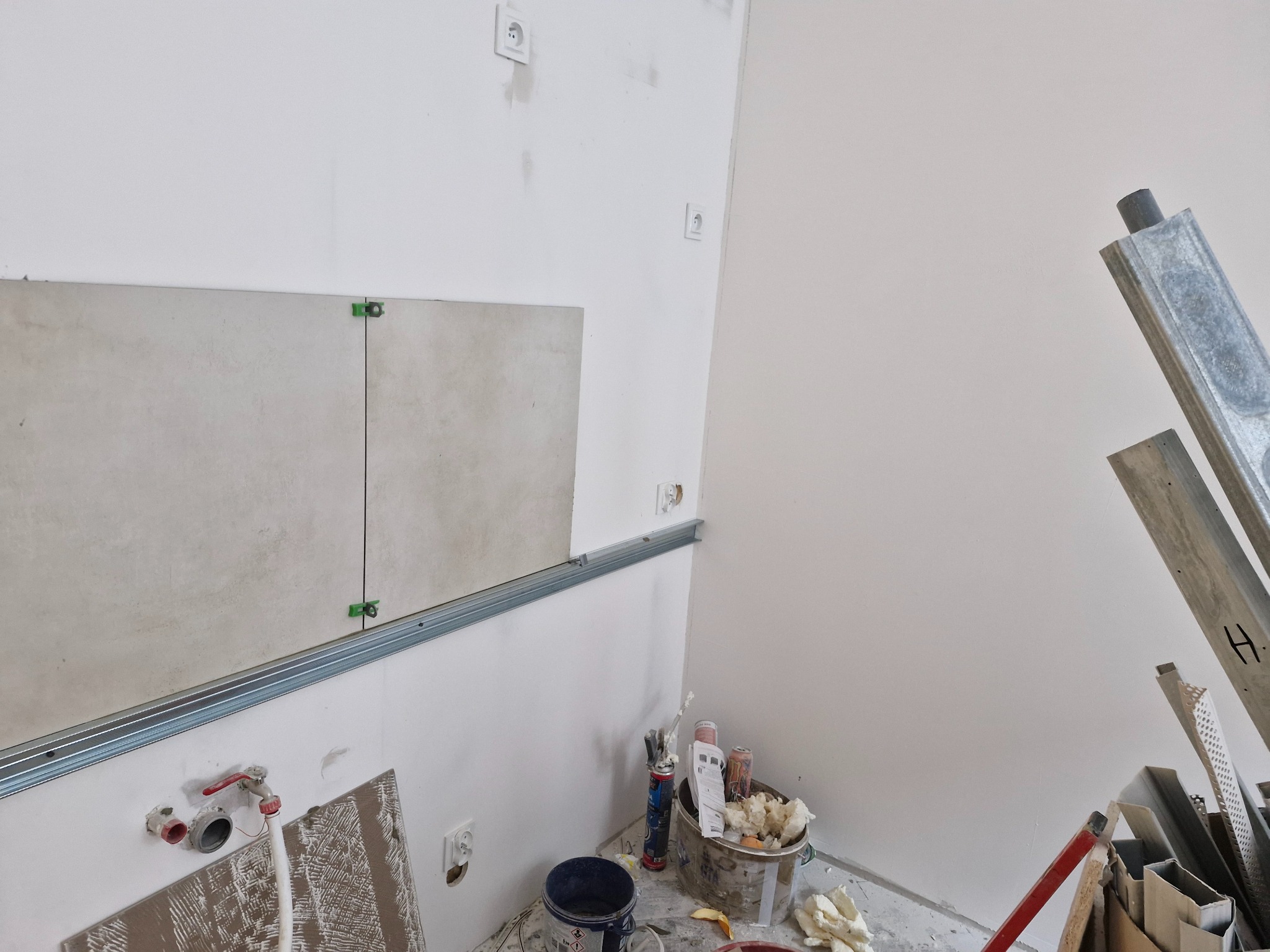 Montaż jasnych płytek na ścianie w łazience, widoczne listwy montażowe, krzyżyki dystansowe, instalacja wodna i akcesoria glazurnicze w wiadrze.