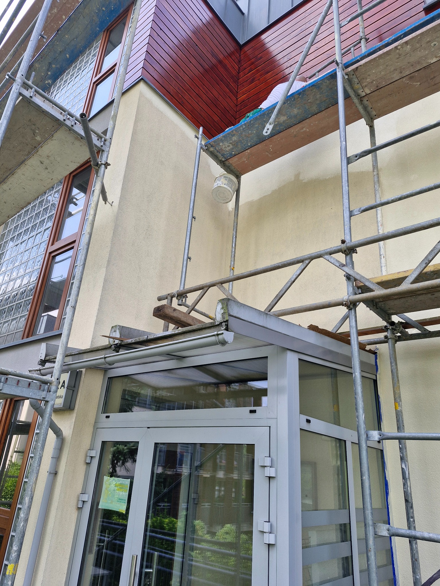 Elewacja budynku z drewnianym wykończeniem poddasza i przeszklonym wejściem, w trakcie malowania, widoczne rusztowania i pracownik na górze.