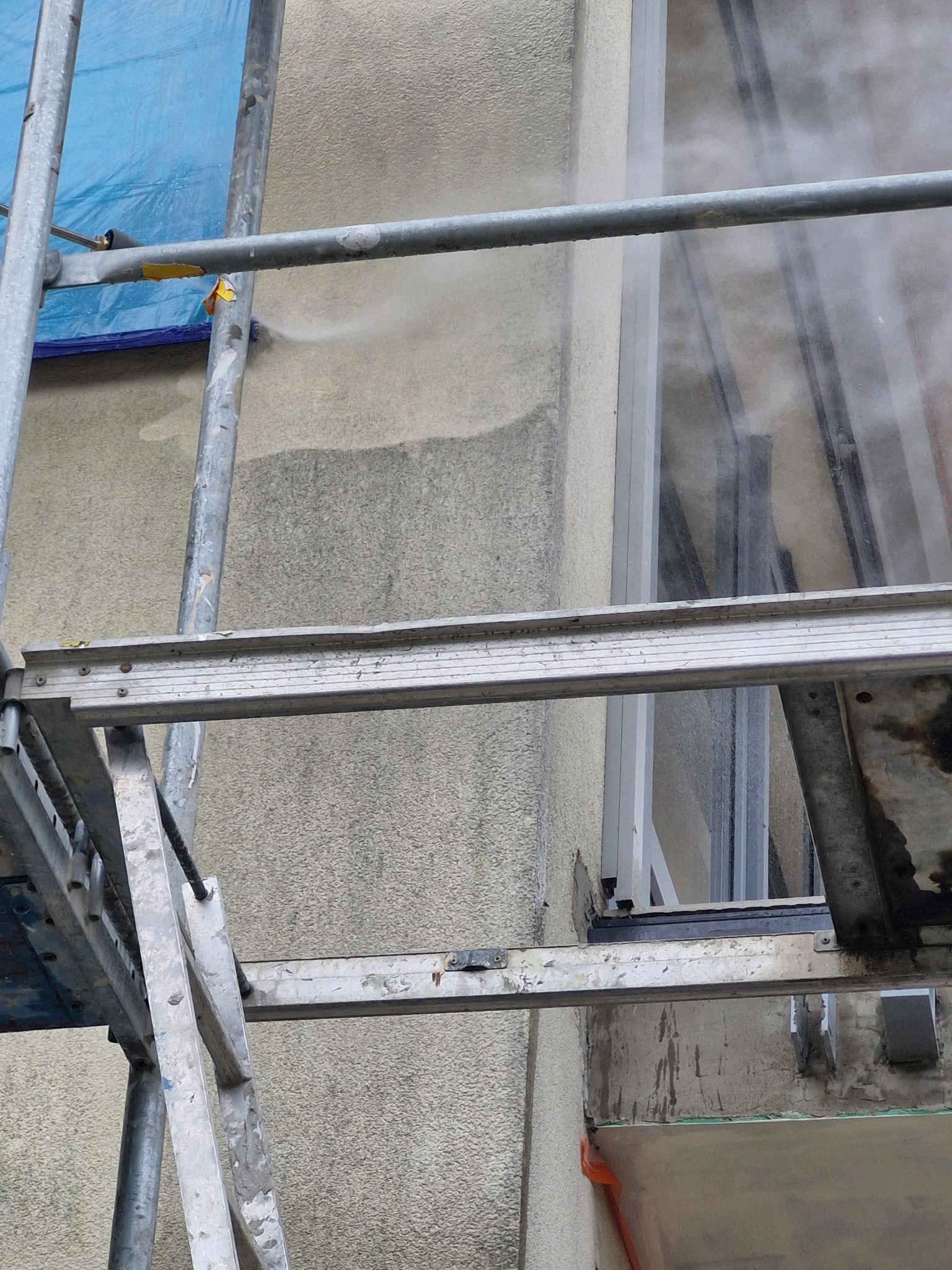 Czyszczenie elewacji budynku strumieniem wody pod ciśnieniem, widoczne rusztowanie i fragment okna.