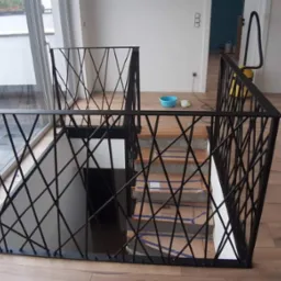 Nowoczesna czarna balustrada metalowa o geometrycznym wzorze krzyżujących się prętów, zabezpieczająca schody wewnętrzne w jasnym pomieszczeniu z drewnianą podłogą, widoczne kartony na stopniach.