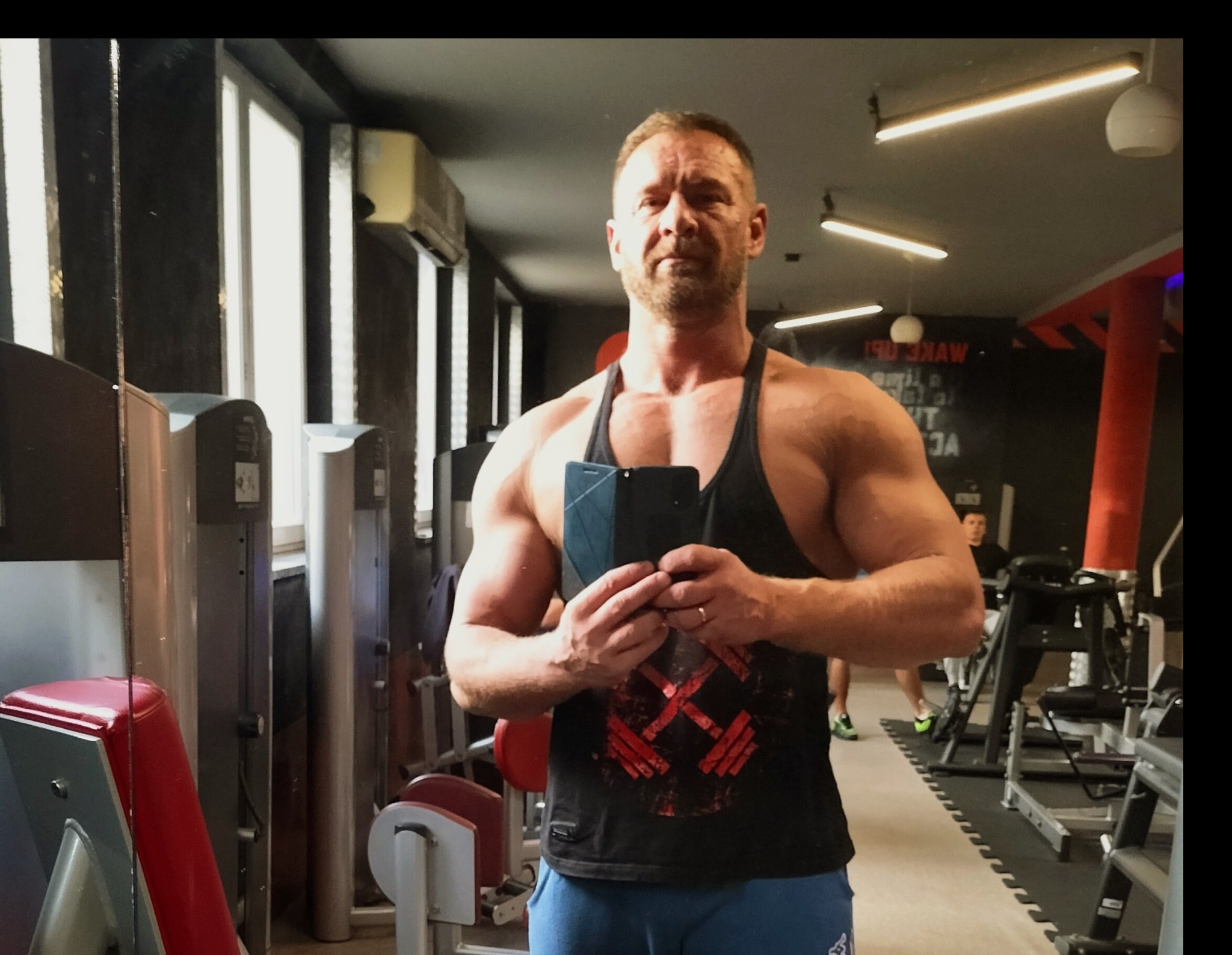 Mężczyzna z wyraźnie zarysowaną muskulaturą, w czarnym podkoszulku z motywem sztangi, robi selfie w siłowni; w tle widoczne maszyny do ćwiczeń i inne osoby.