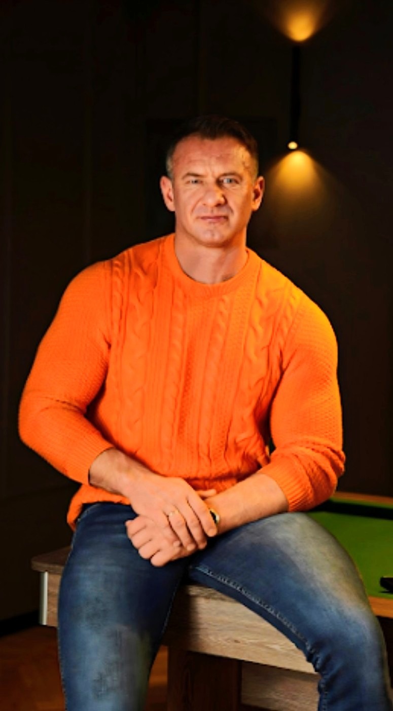 Mężczyzna w pomarańczowym swetrze siedzi na stole bilardowym, patrząc wprost w obiektyw, z oświetleniem punktowym w tle.