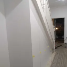 Biała zabudowa pod schodami z widocznymi żółtymi taśmami maskującymi, narzędzia na podłodze sugerują trwający montaż.