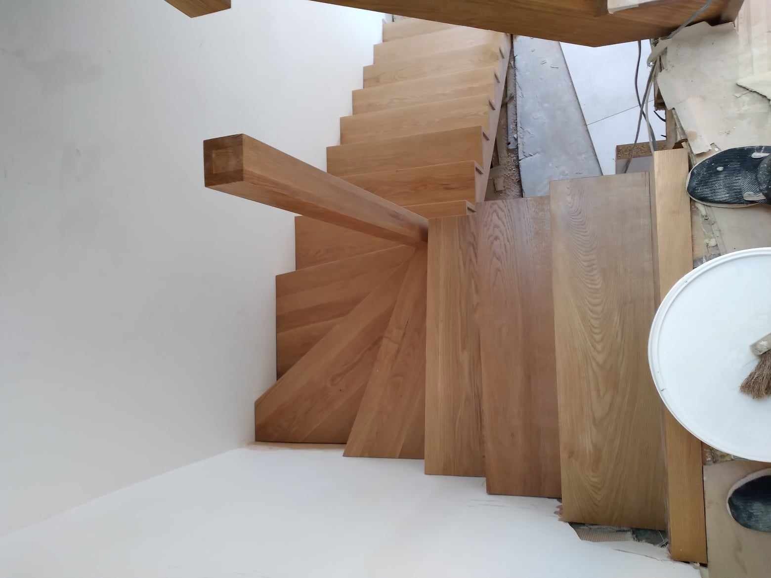 Wysoki kąt widzenia na drewniane schody spiralne z jasnego drewna w trakcie montażu, widoczne niedokończone wykończenia ścian i akcesoria malarskie.