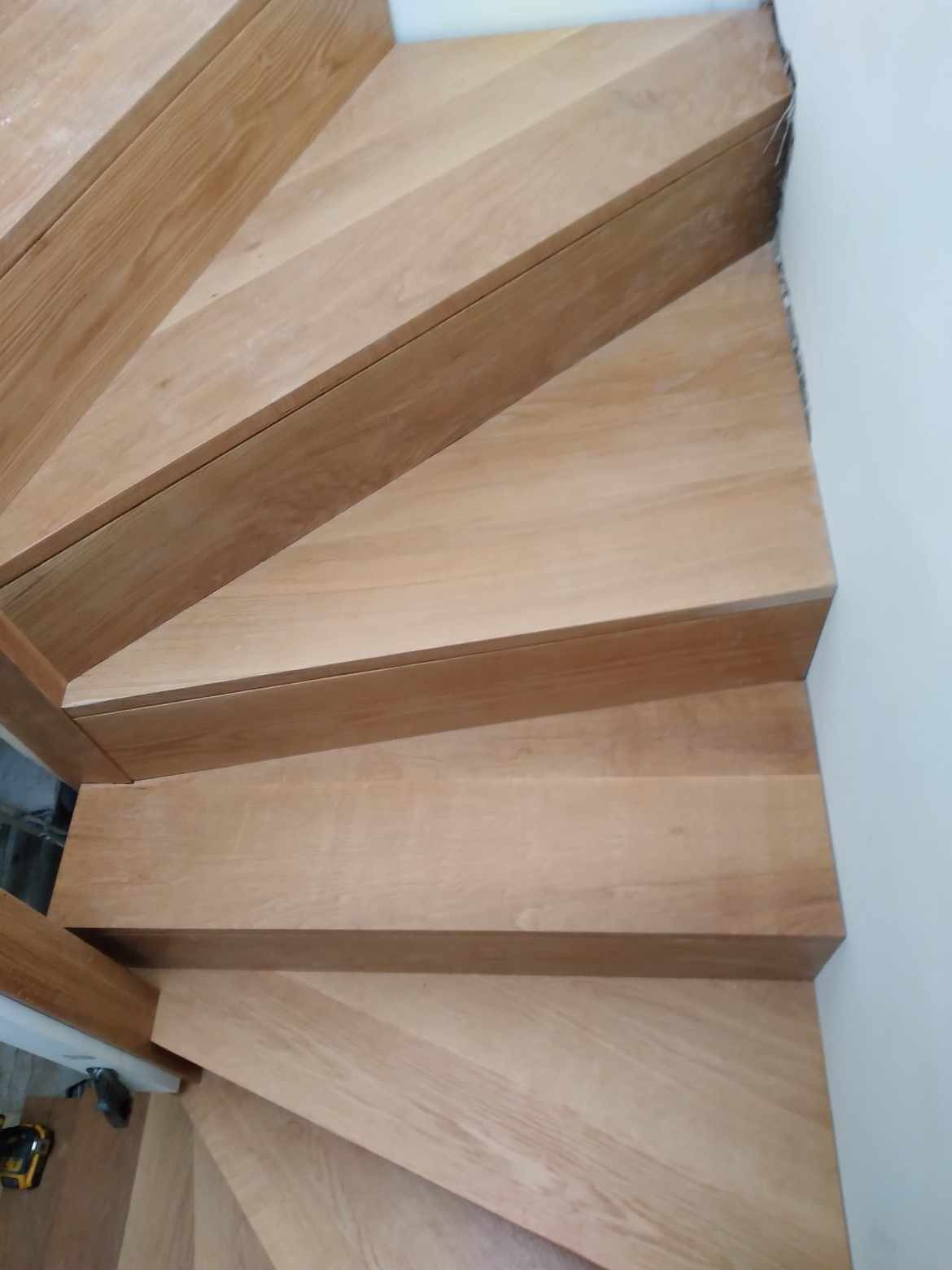 Drewniane schody spiralne z jasnego drewna, widok z góry, z widoczną fakturą drewna i fragmentem białej ściany.