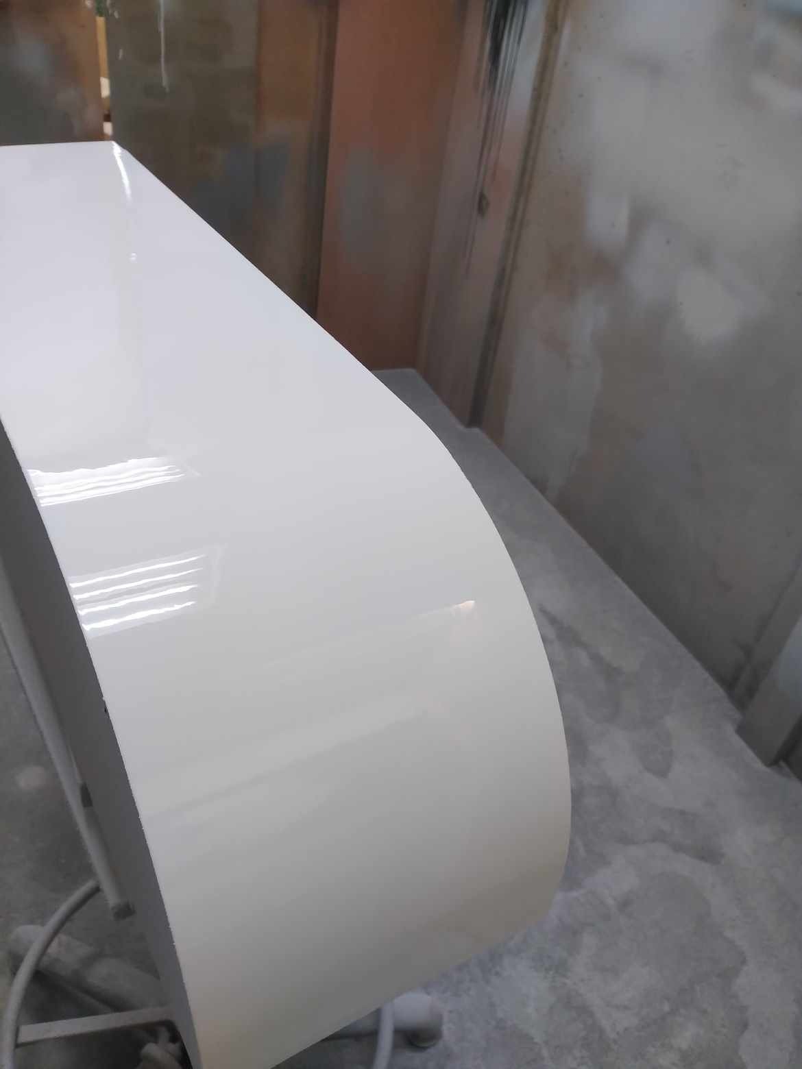 Błyszczący, biały front meblowy z zaokrąglonym brzegiem, prawdopodobnie po lakierowaniu, widoczny w surowym wnętrzu warsztatu.