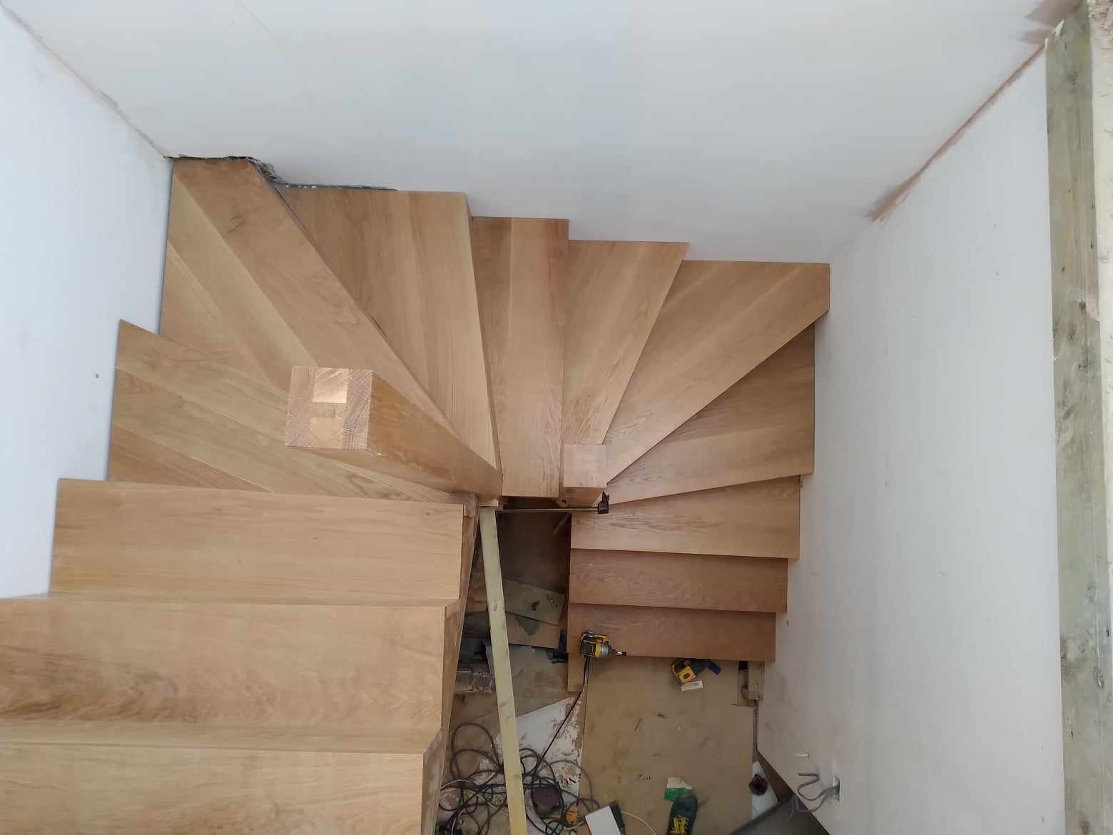 Wewnętrzne schody spiralne z jasnego drewna, widok z góry, w trakcie montażu, widoczne narzędzia i niedokończone wykończenie ścian.