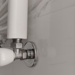 Oleh Khoma - Biały grzejnik łazienkowy z chromowanym zaworem na tle białych płytek imitujących marmur.