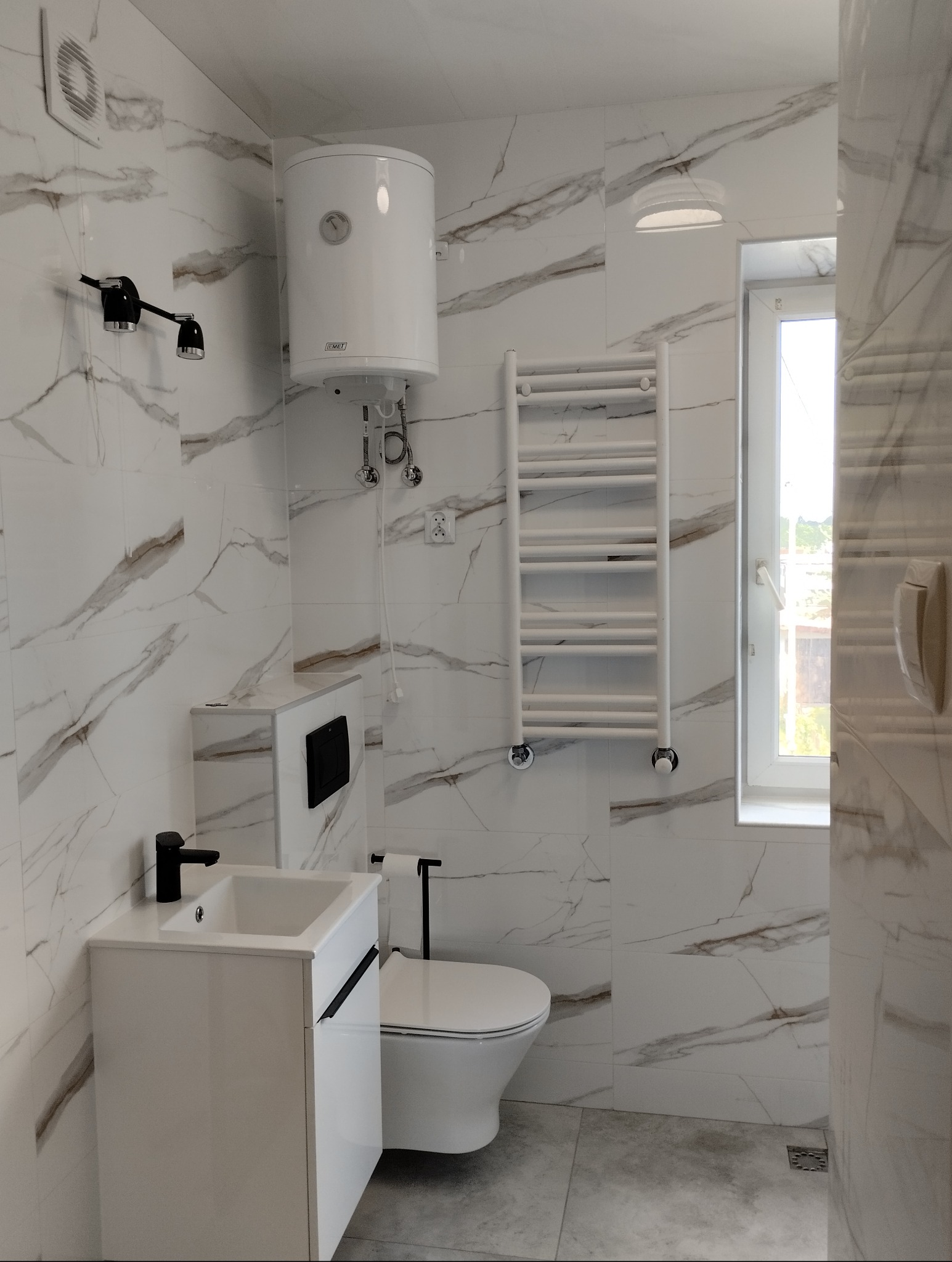 Nowoczesna łazienka z marmurowymi płytkami, białymi meblami, grzejnikiem drabinkowym i podgrzewaczem wody.