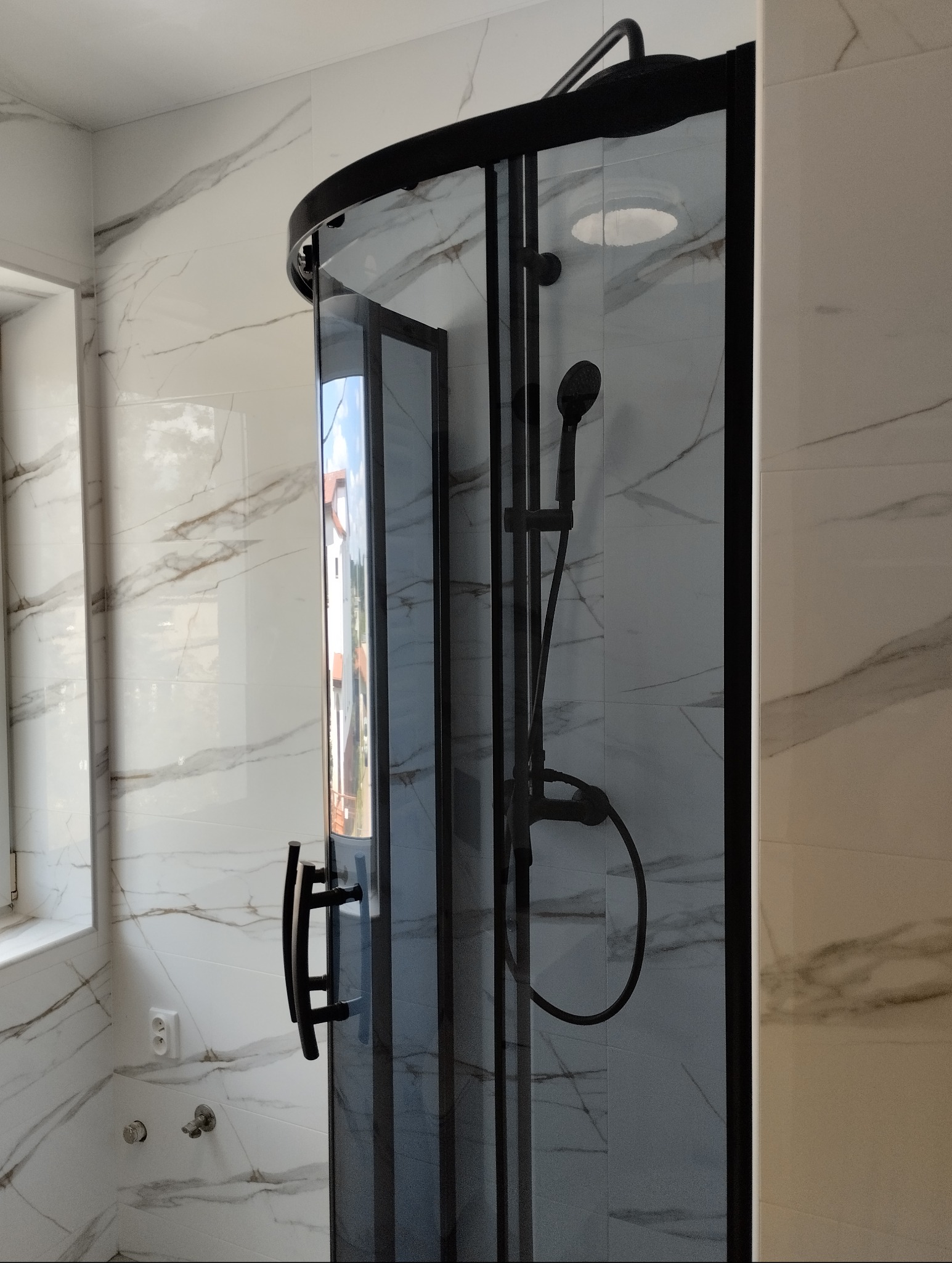 Nowoczesna kabina prysznicowa z czarnymi profilami i armaturą, na tle ściany wyłożonej białymi płytkami imitującymi marmur, widoczne okno z parapetem.