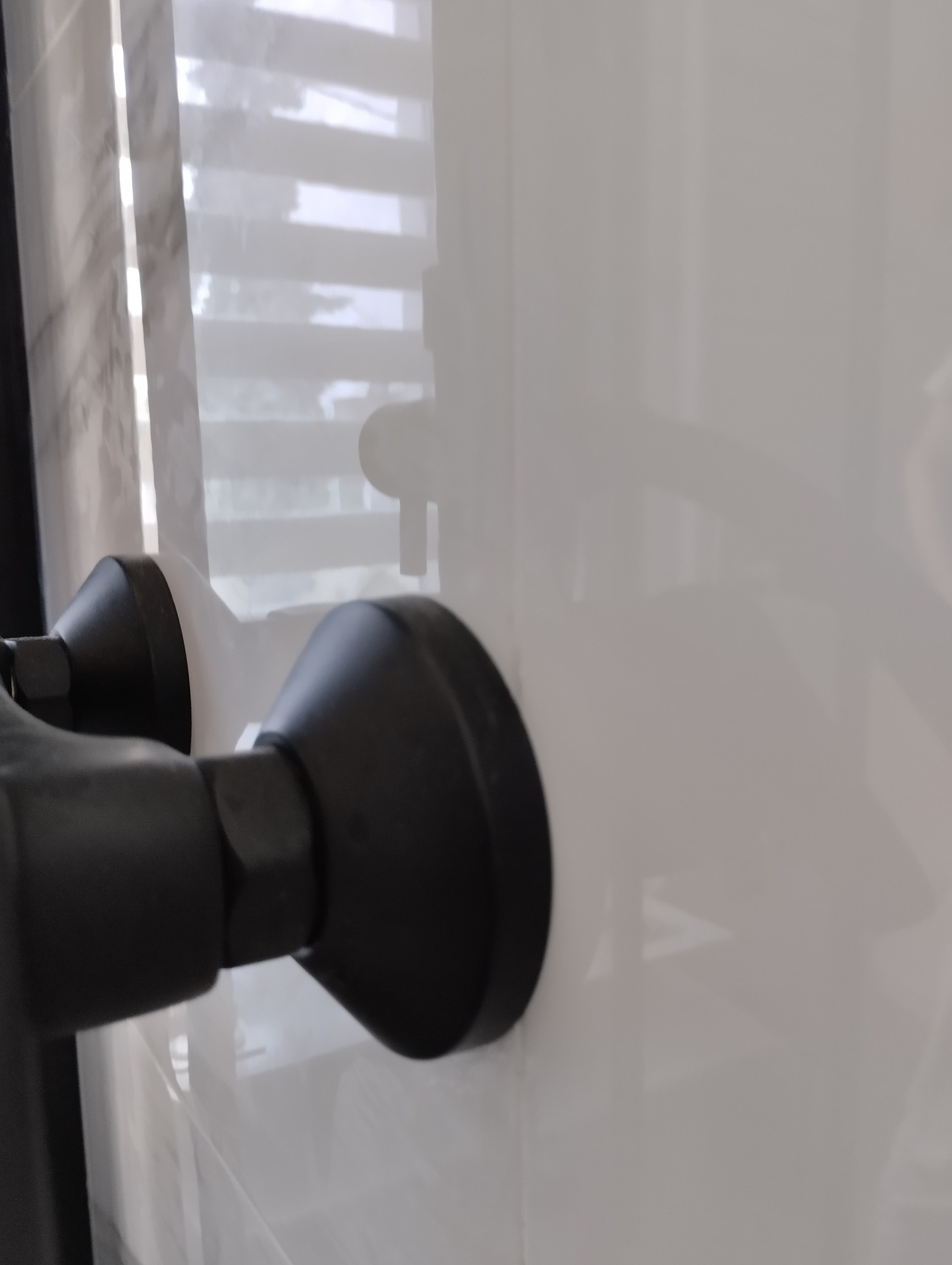 Czarny, nowoczesny kran zamontowany na białej, błyszczącej ścianie w łazience w Łodzi, z widocznymi refleksami światła i żaluzjami w tle.