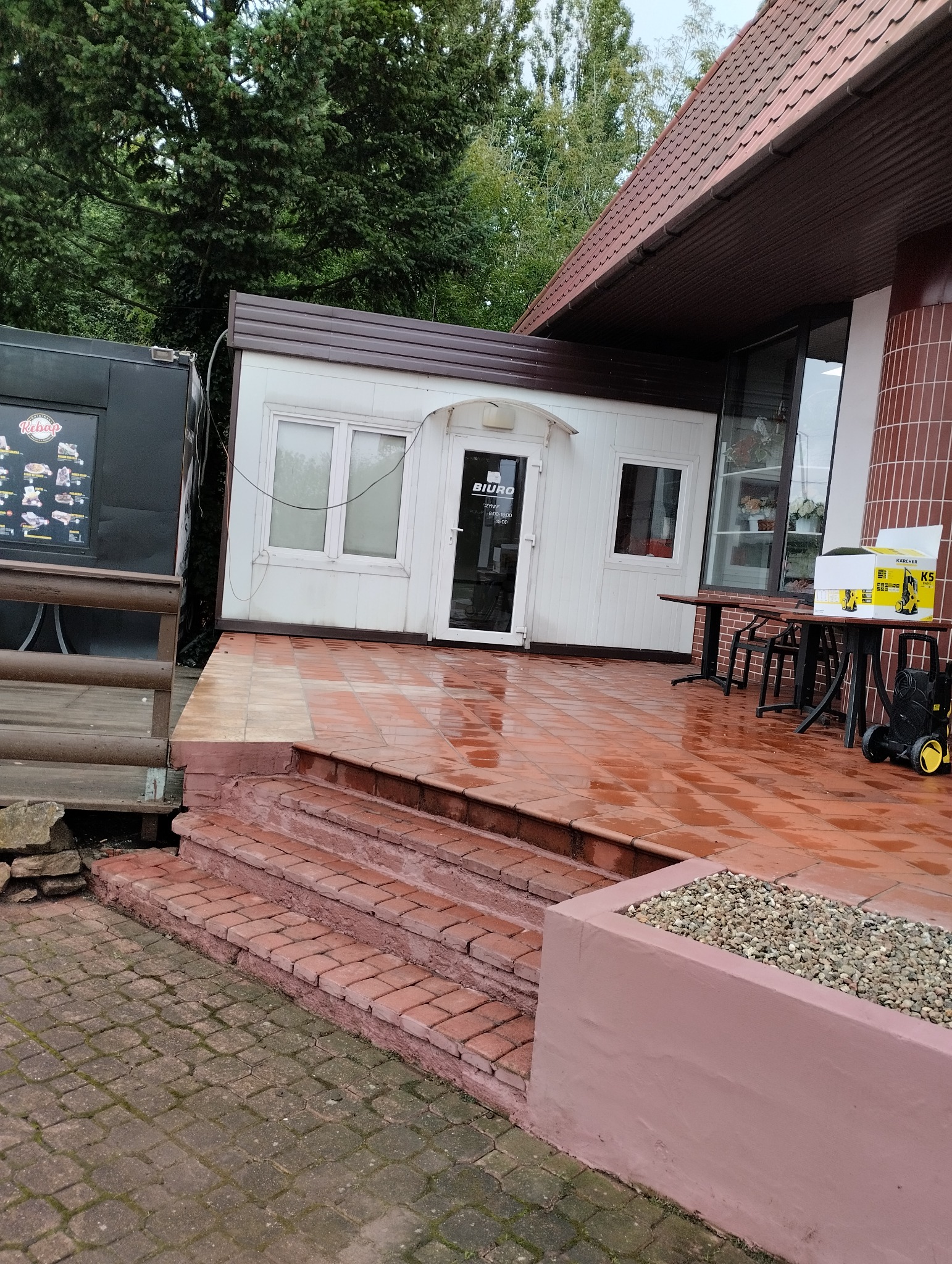 Czerwona, mokra kostka brukowa przed wejściem do biura w kontenerze, obok budka z kebabem, w tle zieleń drzew.