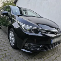 Błyszczący, czarny samochód osobowy marki Toyota, zaparkowany na brukowanym podjeździe przed jasną ścianą budynku.