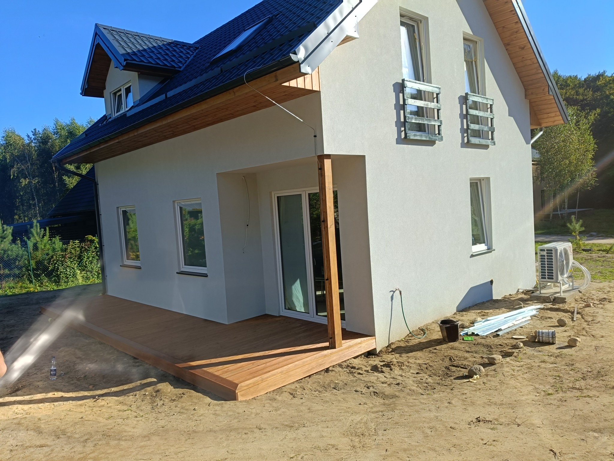 Nowoczesny, dwukondygnacyjny dom z drewnianym tarasem w trakcie budowy. Widoczne elementy wykończeniowe, klimatyzator i narzędzia na posesji. Jasne tynki i ciemna dachówka.