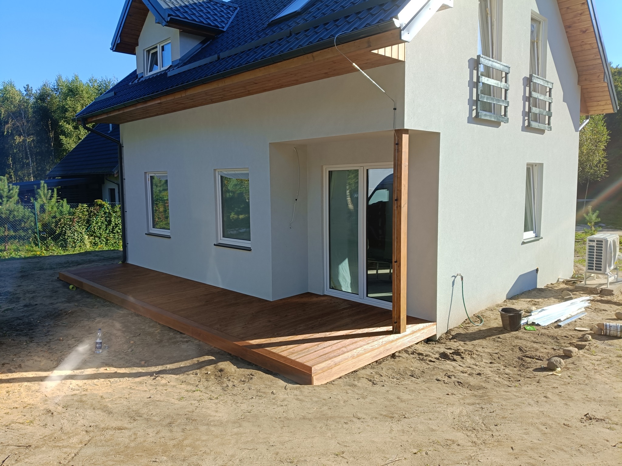 Nowoczesny dom z drewnianym tarasem w Szymbarku. Jasna elewacja, grafitowy dach, okna z białymi ramami. Widoczny fragment ogrodu i butelka z wodą na ziemi.