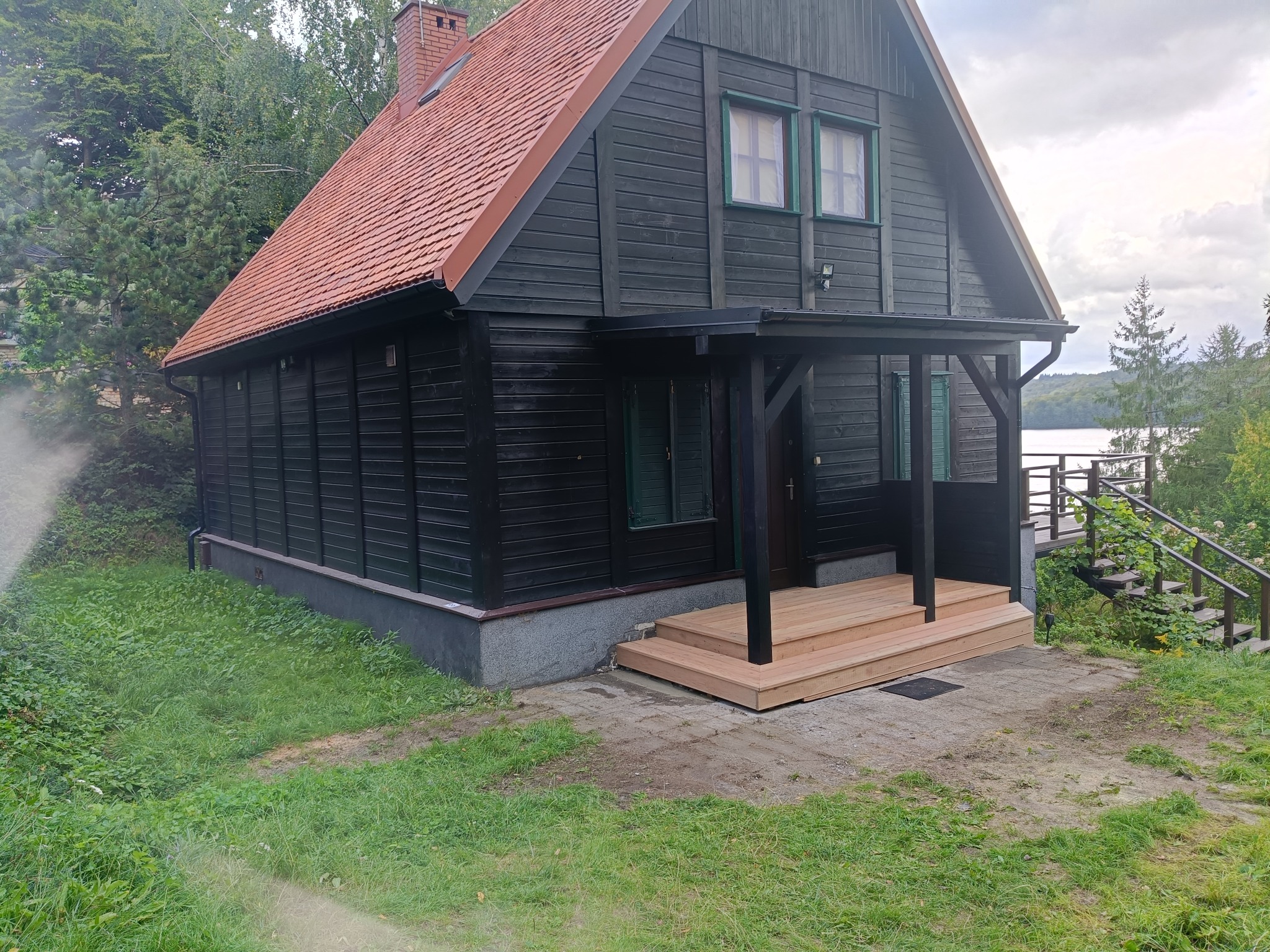 Czarny dom szkieletowy z czerwonym dachem, drewnianym gankiem i zielonymi oknami, otoczony zielenią, z widokiem na jezioro w tle.