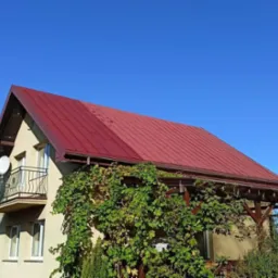 Czerwony, metalowy dach domu jednorodzinnego po malowaniu, widoczny fragment elewacji z balkonem i zarośniętą pergolą na tle błękitnego nieba.