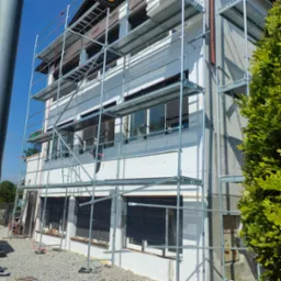 Elewacja domu w trakcie ocieplania styropianem, widoczne rusztowania, okna z roletami zewnętrznymi i fragment starej, szarej elewacji z rurą spustową.