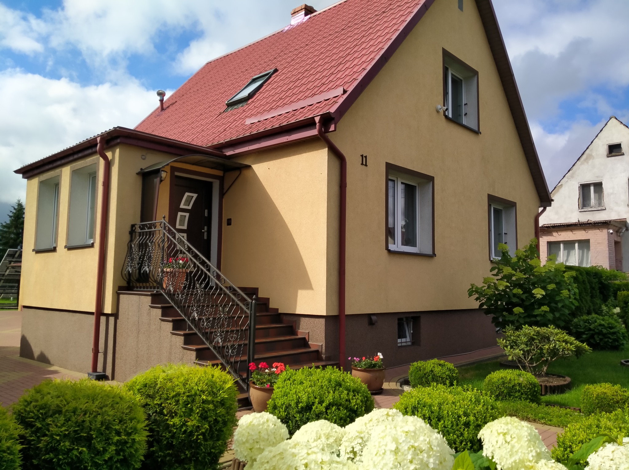 Dom jednorodzinny z elewacją w odcieniach beżu, czerwonym dachem i zadbanym ogrodem z kwiatami w donicach oraz ozdobnymi krzewami.