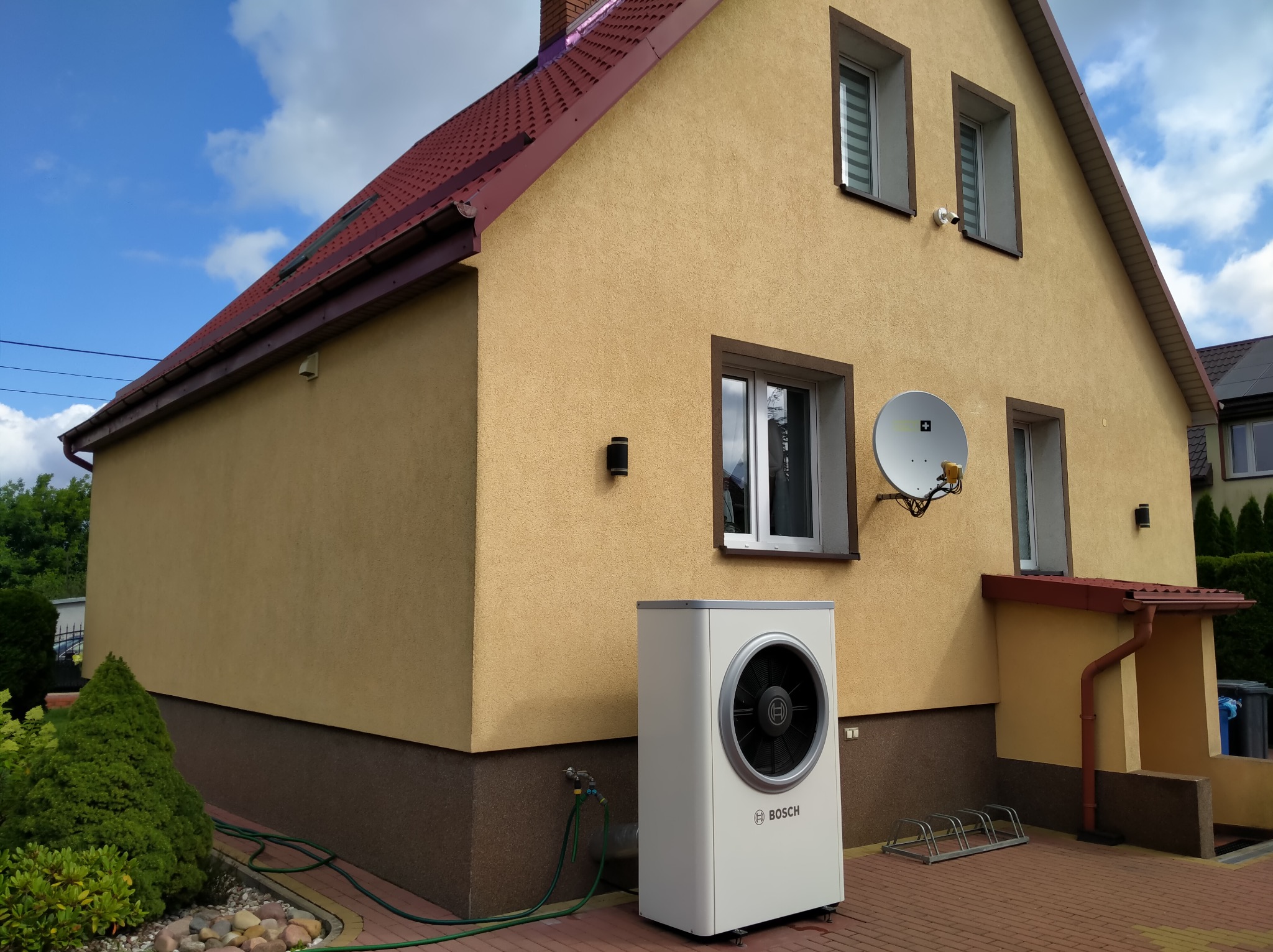 Żółta elewacja domu jednorodzinnego z jasnobrązowymi obramowaniami okien i nową pompą ciepła Bosch, widoczna z boku, w tle błękitne niebo z chmurami.