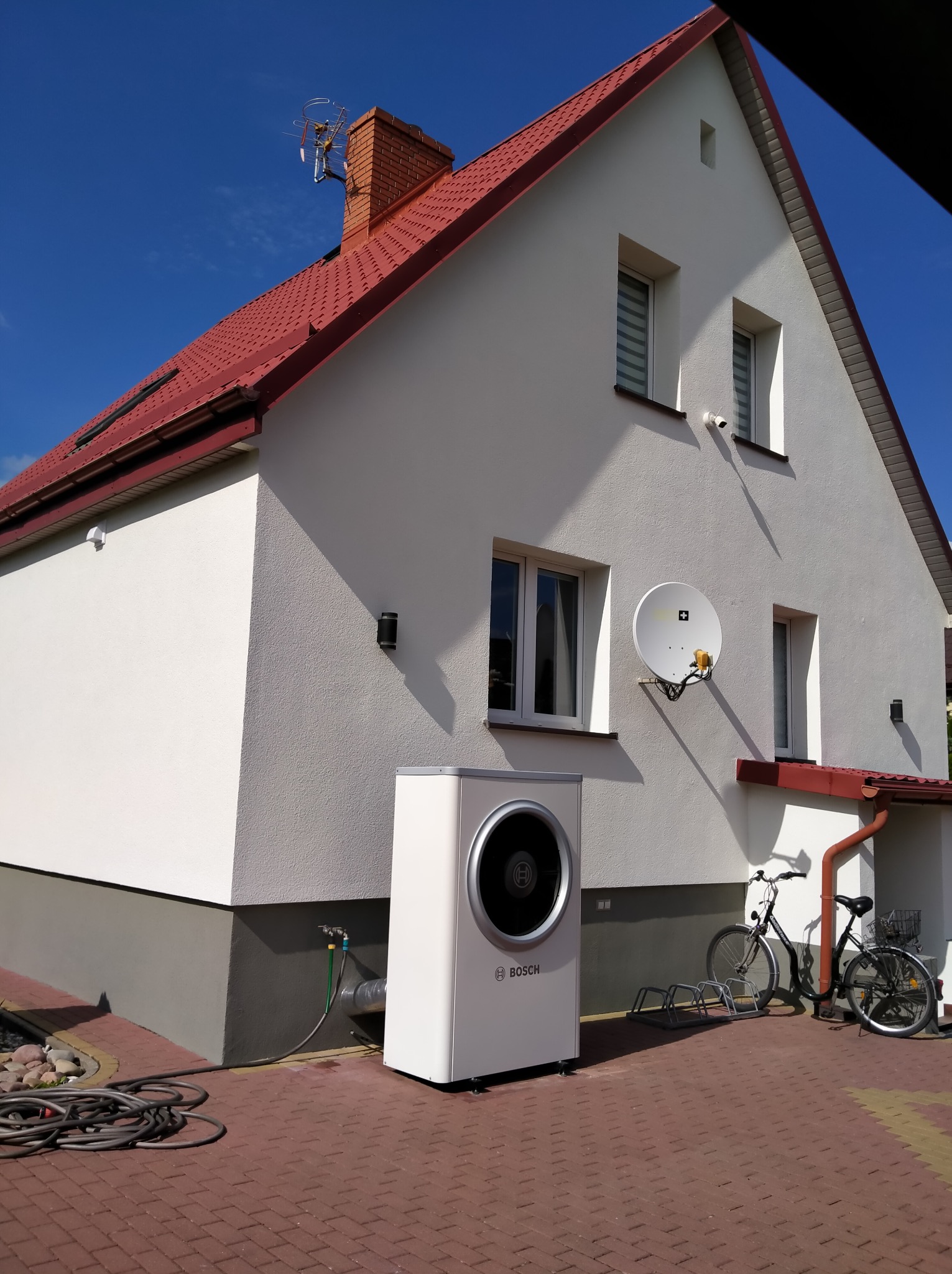 Biały dom jednorodzinny z czerwoną dachówką, na elewacji zamontowana antena satelitarna i lampy zewnętrzne, obok domu pompa ciepła marki Bosch, dwa rowery i zwinięty wąż ogrodowy.