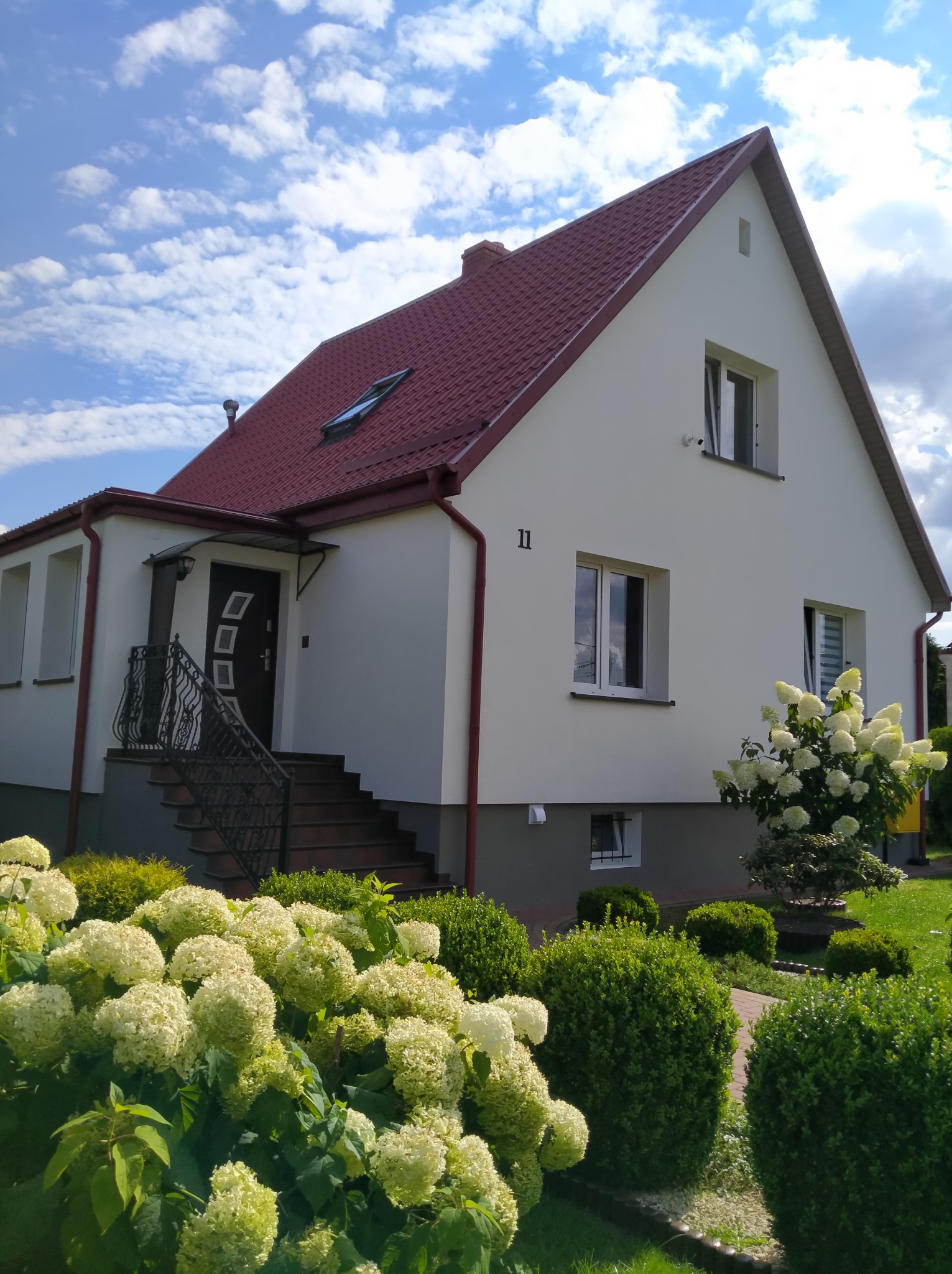 Jasna elewacja domu jednorodzinnego z czerwonym dachem, numerem 11 i bujnymi hortensjami w ogrodzie, widok z poziomu ogrodu w słoneczny dzień.