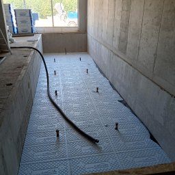 MIX-MASTERS KONRAD SZKODA - Przygotowanie podłoża pod wylewkę: białe płyty styropianowe z nadrukiem Hydro Styromax ułożone na dnie betonowego pomieszczenia, widoczny wąż i wystające pionowe elementy, w tle okno z widokiem...