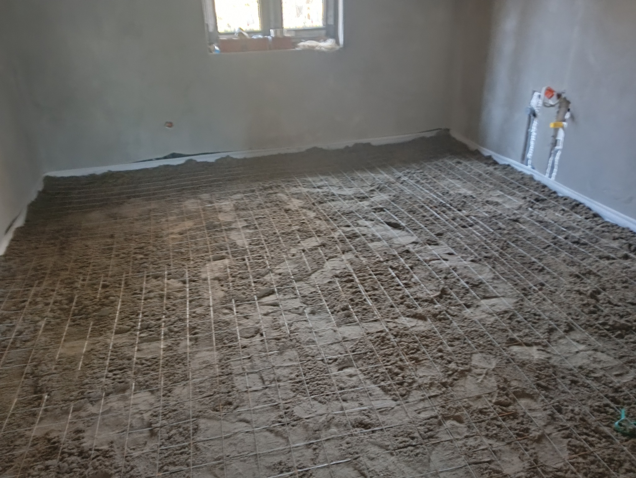 Przygotowanie podłoża pod wylewkę w pomieszczeniu z oknem, widoczna siatka zbrojeniowa na warstwie izolacji przy ścianach.