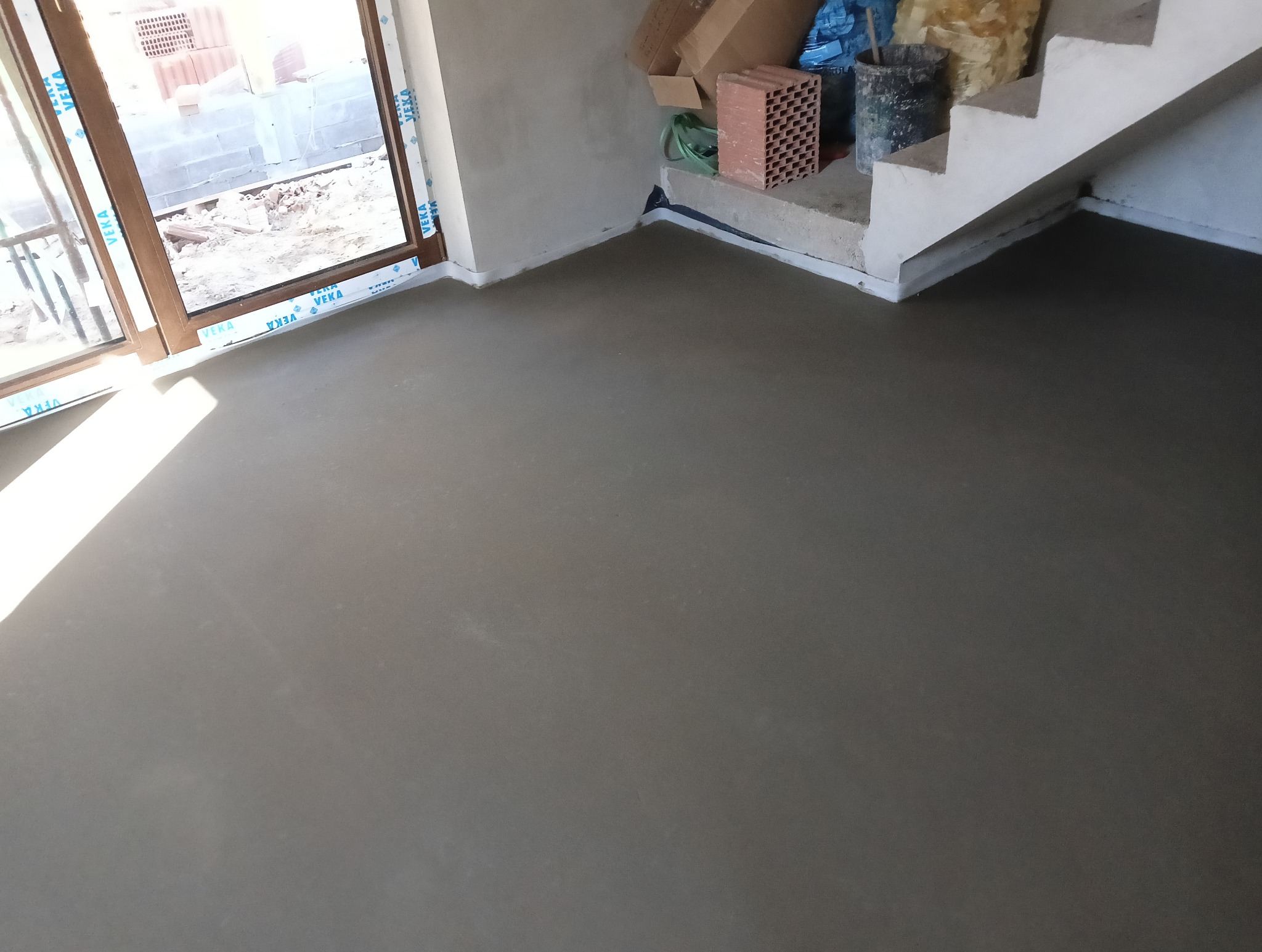 Świeżo wylana, gładka powierzchnia podłogi w surowym wnętrzu z widocznymi schodami i oknem z nieotynkowanym murem na zewnątrz.