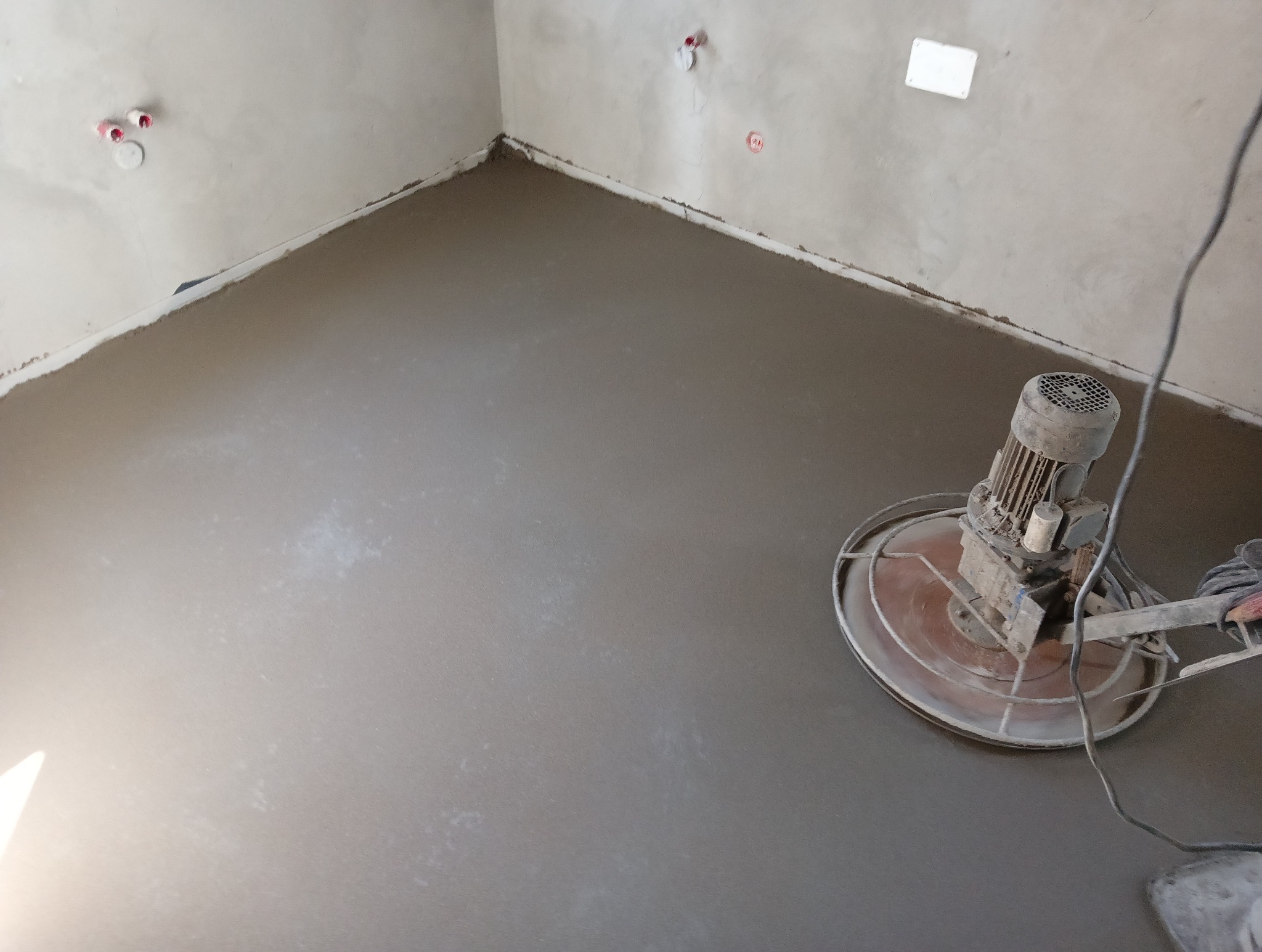 Świeżo wylana, szara posadzka wyrównywana maszyną z obrotowym talerzem w surowym wnętrzu.