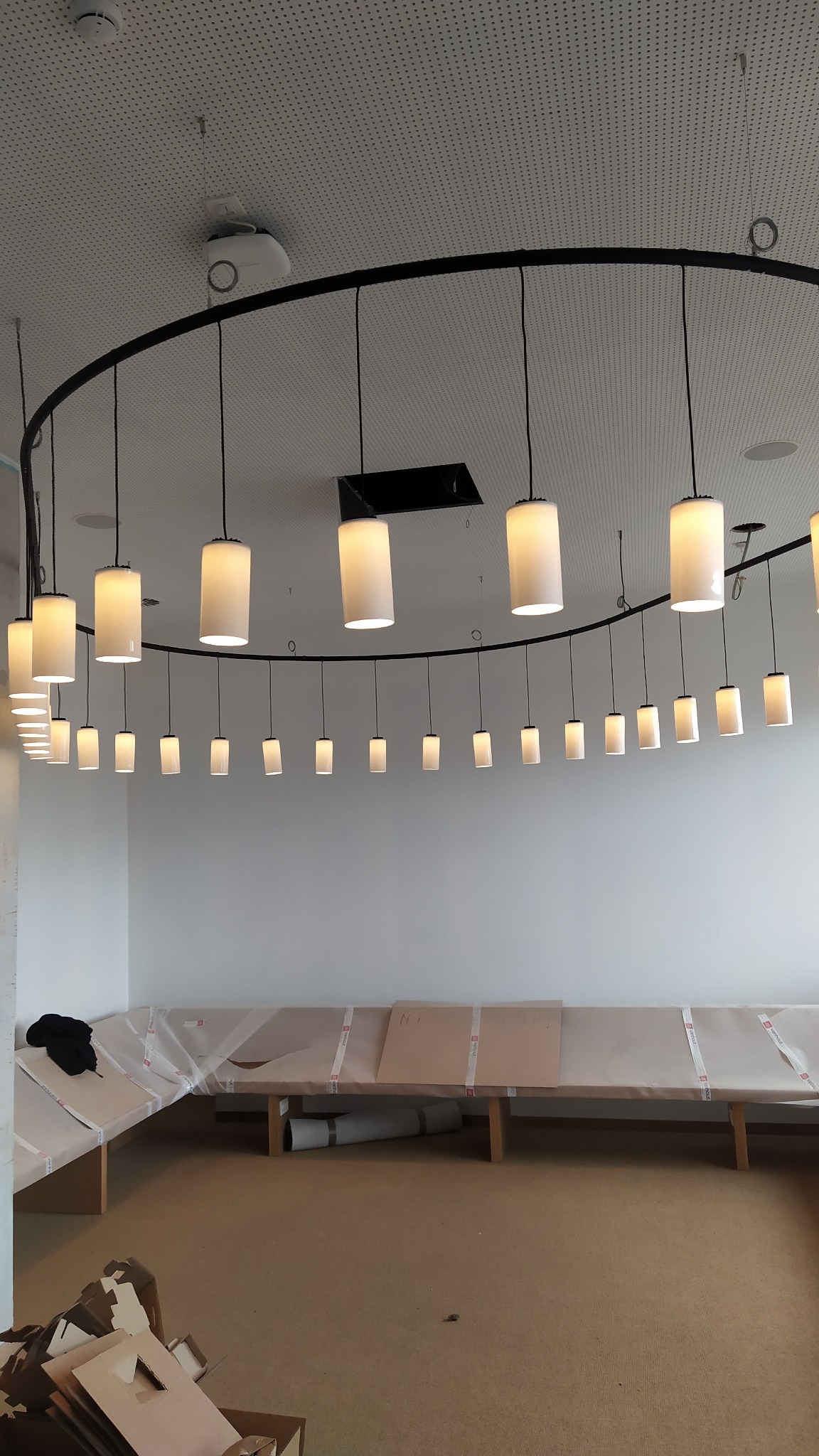Okrągła instalacja oświetleniowa z cylindrycznymi lampami w biurze, widoczne meble zabezpieczone folią ochronną, sufit podwieszany z perforacją.