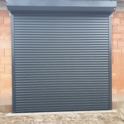 Admar Monika Markowska - Zamknięta, grafitowa roleta zewnętrzna na tle surowej ściany z cegły dziurawki. Widoczne przewody elektryczne. Ujęcie z dołu, podkreślające solidność konstrukcji.