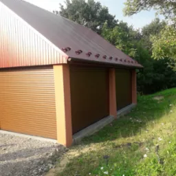 Dwa brązowe garaże z rolowanymi bramami, brązowy metalowy dach, widok z zewnątrz, trawa i drzewa w tle.