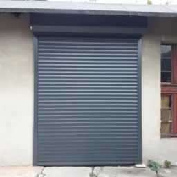 Zamknięta, szara roleta zewnętrzna zamontowana na szarej ścianie budynku z widocznym fragmentem okna z drewnianą ramą.