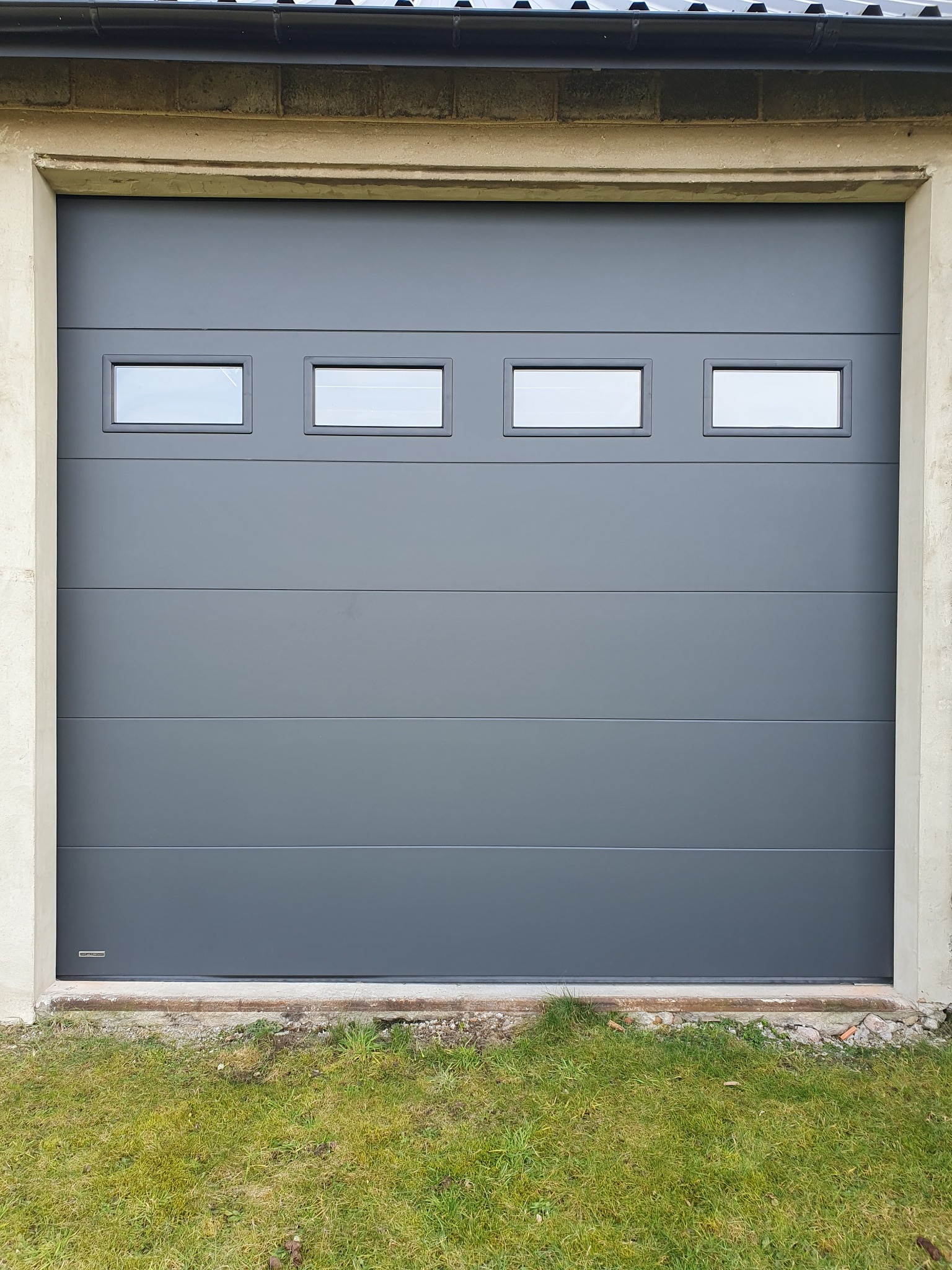 Szara segmentowa brama garażowa z czterema prostokątnymi okienkami, zamontowana w betonowym otworze, z widoczną trawą u dołu. Nowoczesny design.