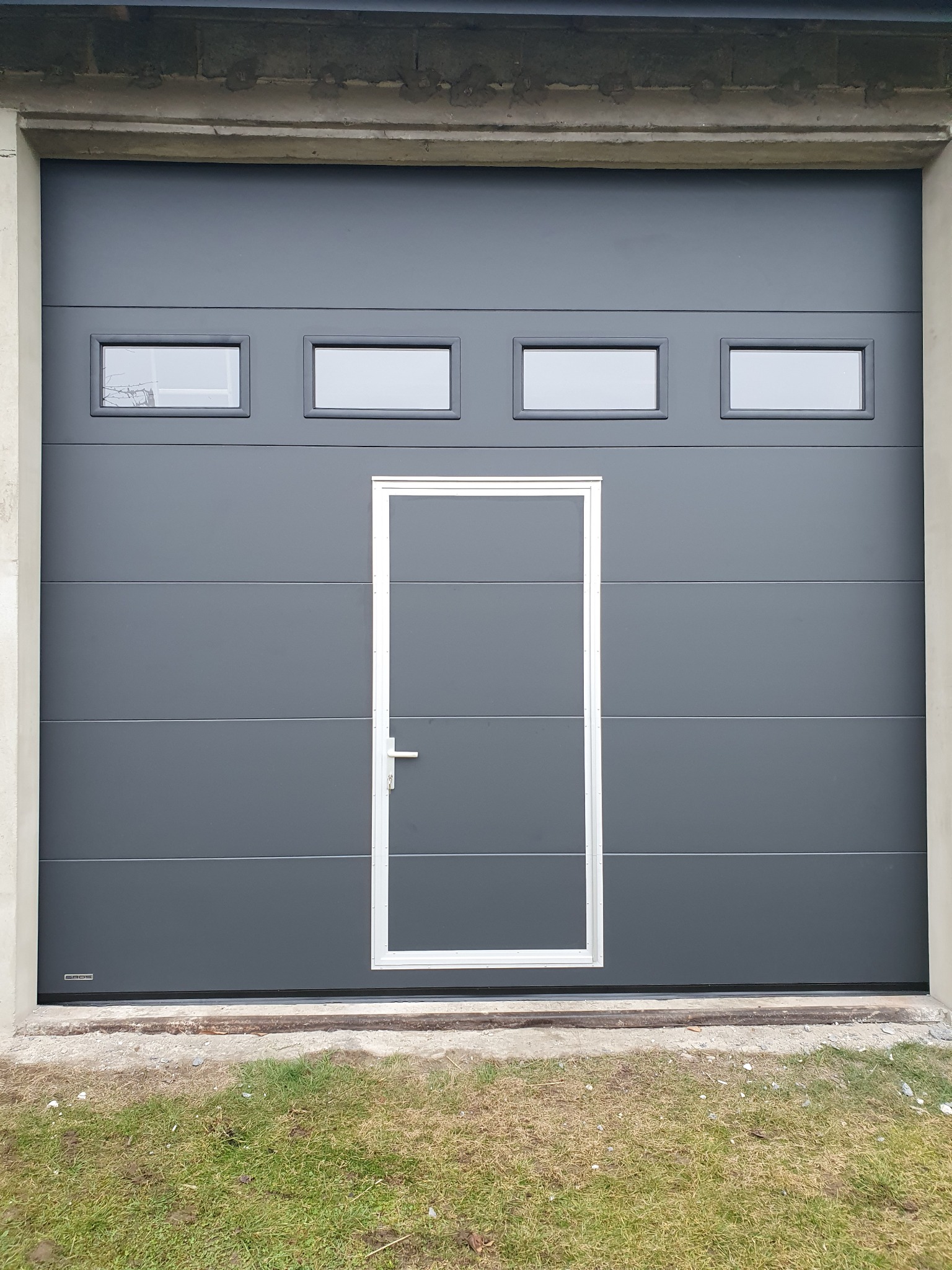 Szara segmentowa brama garażowa z czterema małymi oknami u góry i drzwiami wejściowymi pośrodku, zamontowana w betonowym otworze, z widoczną trawą u dołu.