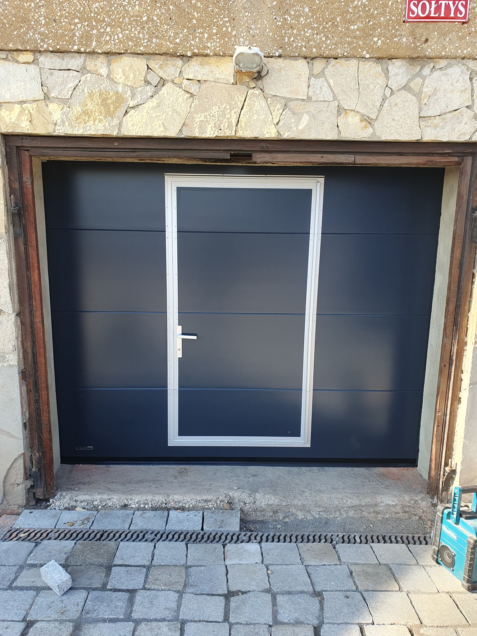 Ciemna brama garażowa segmentowa z drzwiami wejściowymi pośrodku, zamontowana w kamiennym budynku. Widoczny fragment kostki brukowej i narzędzie.