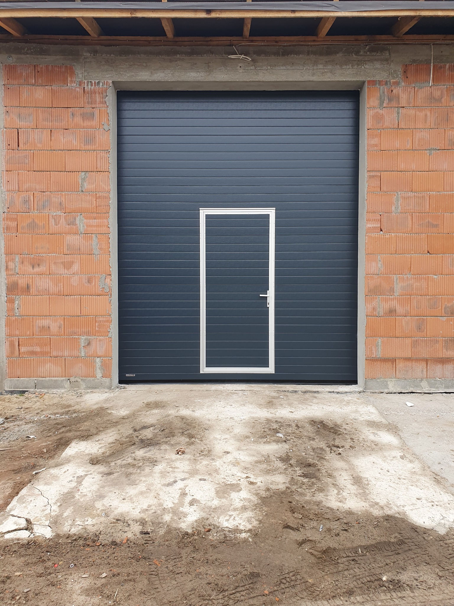 Ciemnoszara brama garażowa segmentowa z drzwiami wejściowymi, zamontowana w surowym murze z cegły. Widoczne ślady budowy na posadzce.
